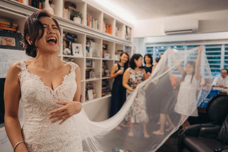 Bride laughing joyfully during wedding prep in Singapore, with bridesmaids holding veil.