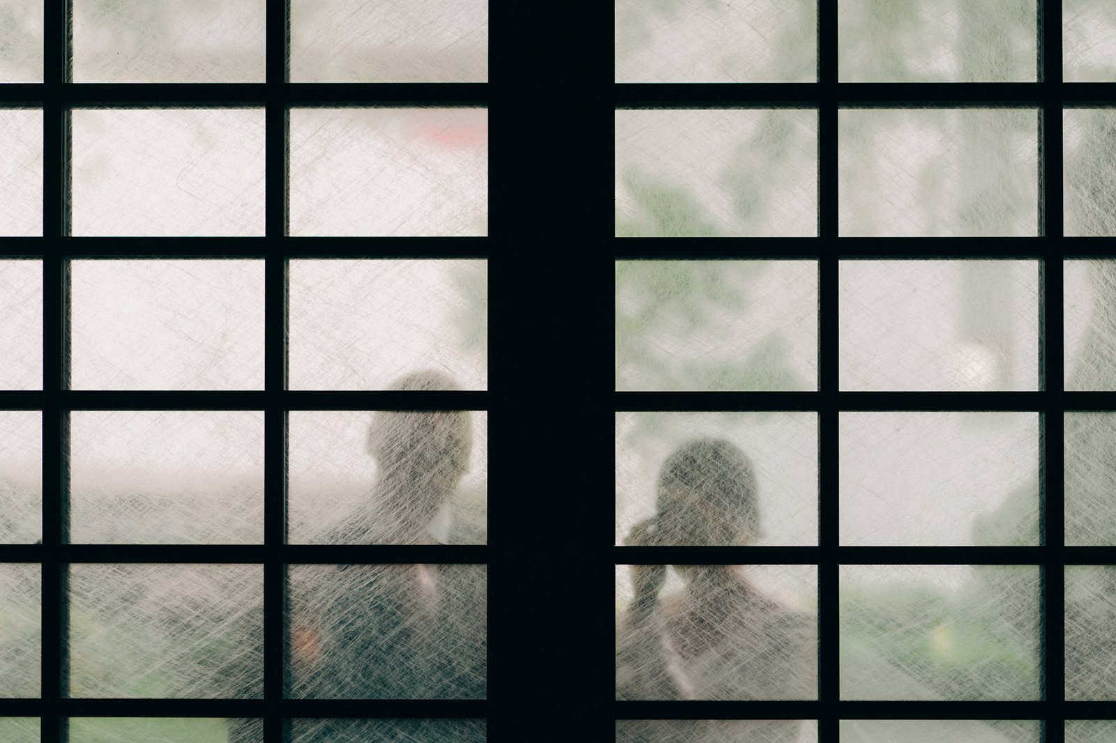 Two figures are silhouetted behind a frosted glass window divided into a grid, suggesting a private moment or a glimpse into another space.