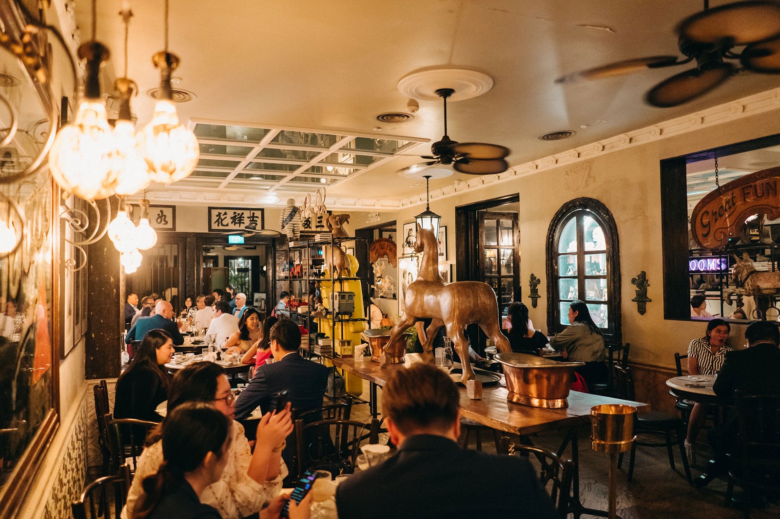Elegant restaurant interior at The English House, Singapore, with guests dining at tables, illuminated by vintage pendant lights, featuring a striking horse statue and ornate decor.
