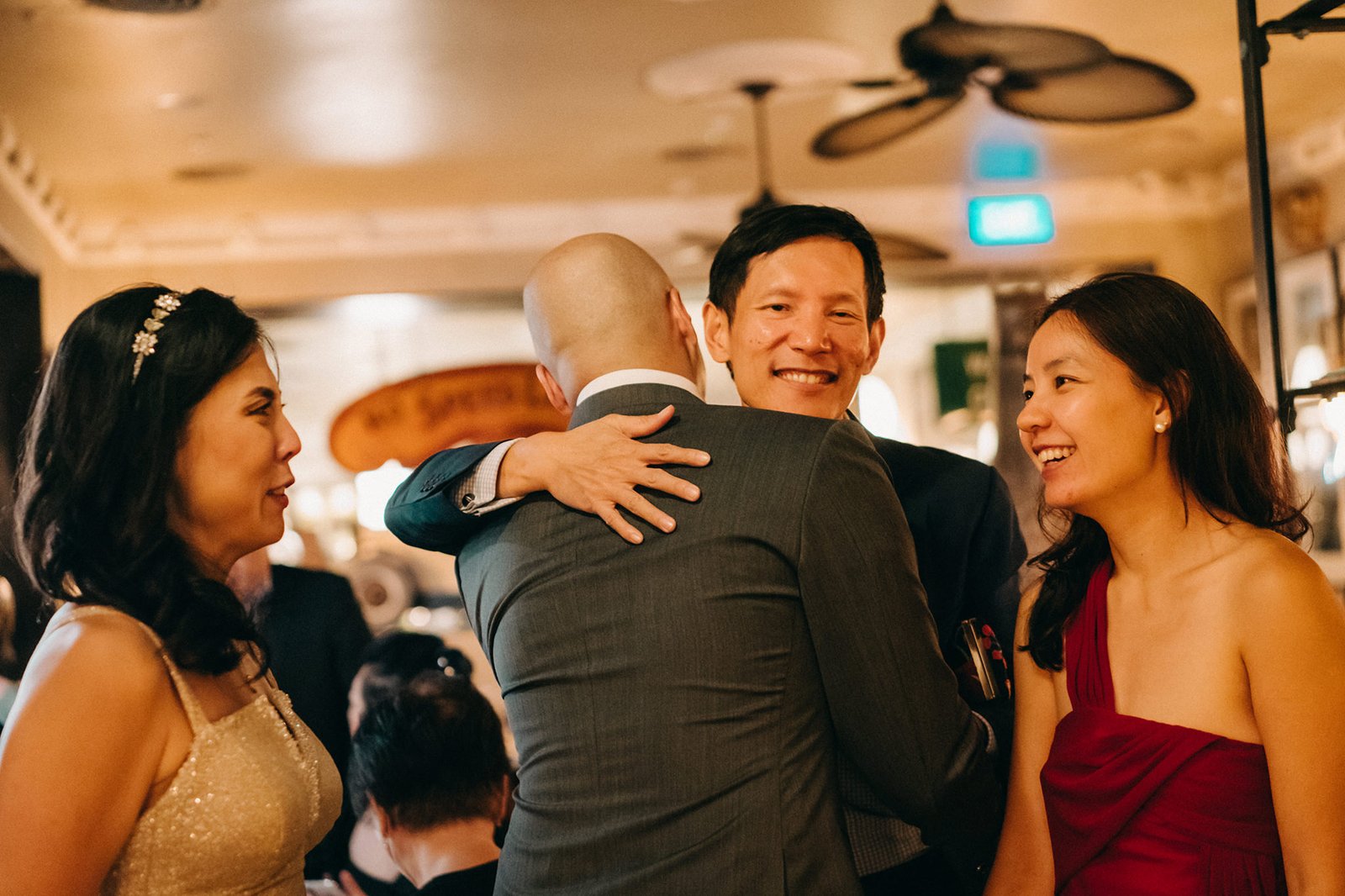 A warm and joyful moment at The English House wedding in Singapore, as a man in a grey suit embraces a friend, while the bride in a red dress and a guest in a gold dress smile nearby.