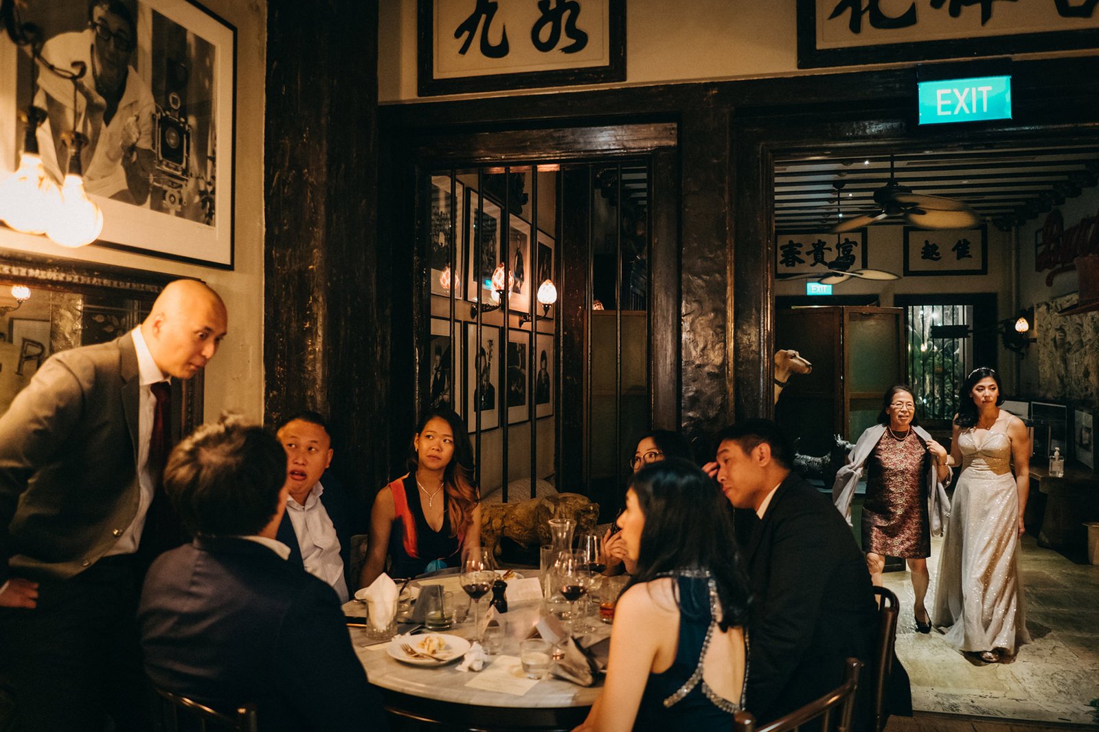 A wedding reception at The English House Singapore, with guests seated at a round table, enjoying a meal and conversation. The elegant atmosphere features vintage decor and framed photographs.