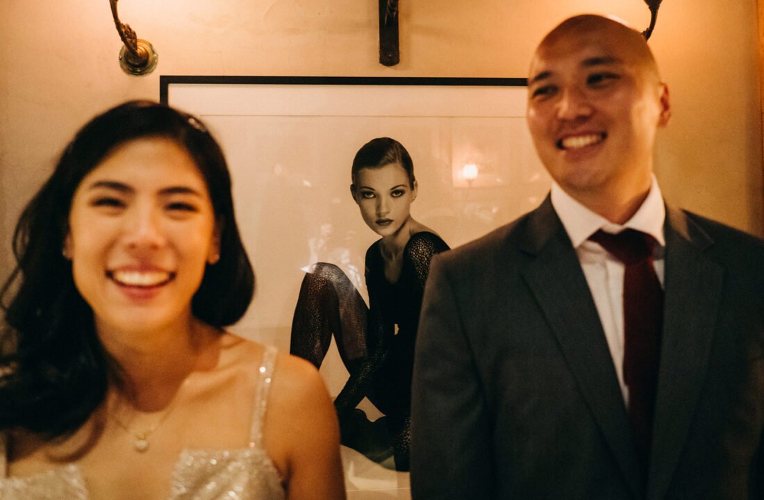 A smiling couple, Kin and Christine, pose in front of a framed photo of Kate Moss at The English House Wedding Singapore.