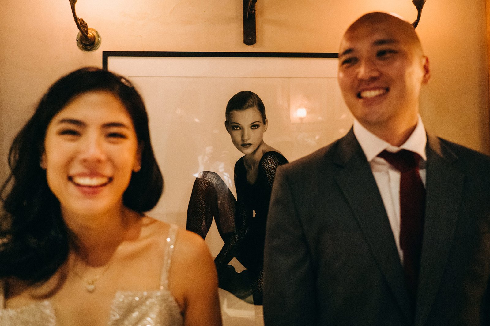 A smiling couple, Kin and Christine, pose in front of a framed photo of Kate Moss at The English House Wedding Singapore.
