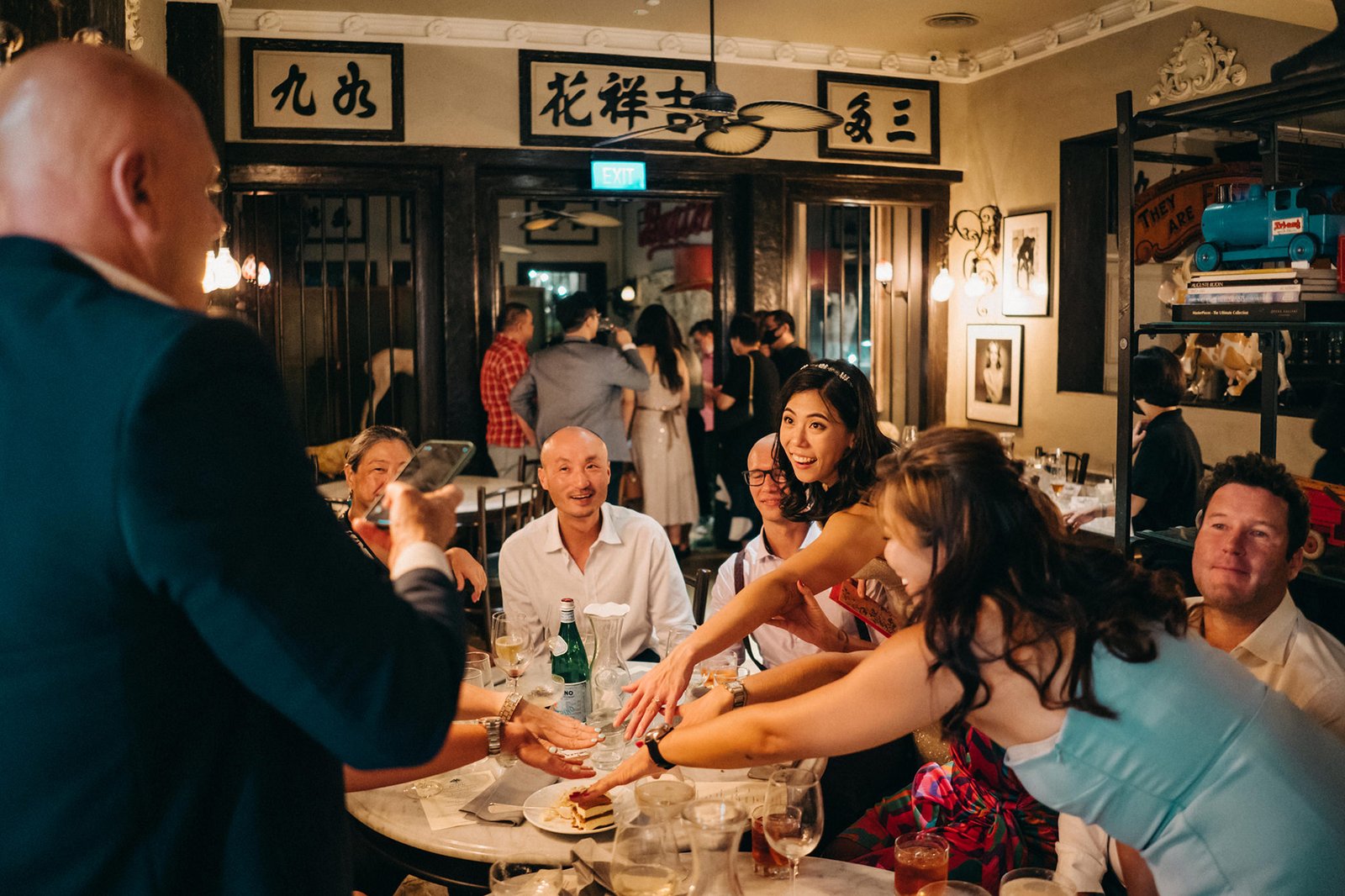 A lively wedding reception at The English House Wedding Singapore, with guests seated at a round table, enjoying a meal and interacting. A bride in a white dress smiles joyfully.