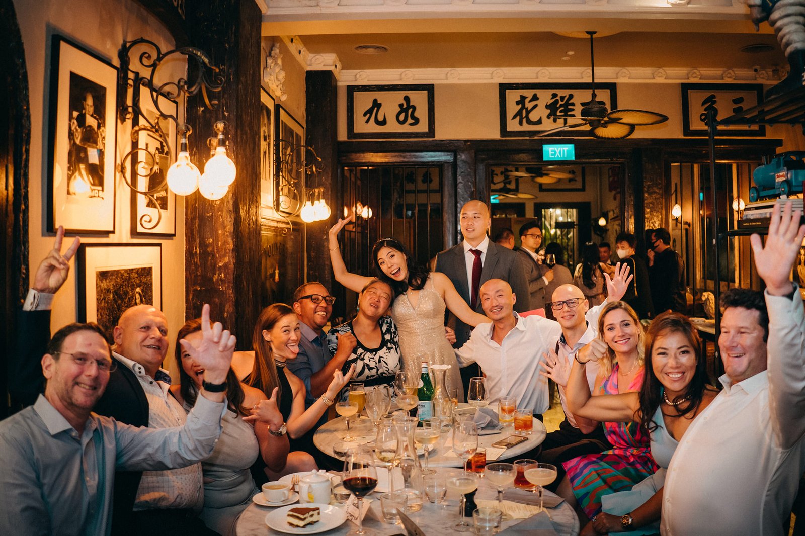 A joyful wedding party poses for a photo at The English House Wedding Singapore, with the bride in a gold dress and groom in a suit, surrounded by friends and family in a dimly lit, elegant restaurant.