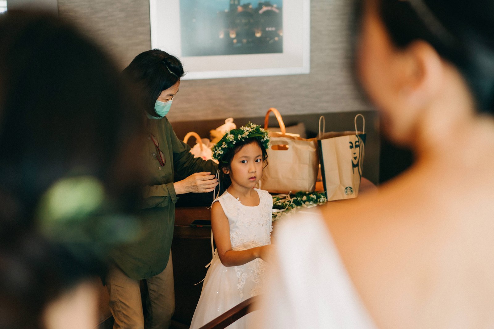 A young flower girl with a flower crown looks at the camera while a woman in a mask adjusts her dress before a wedding at the Keyaki Garden Pavilion in Singapore.