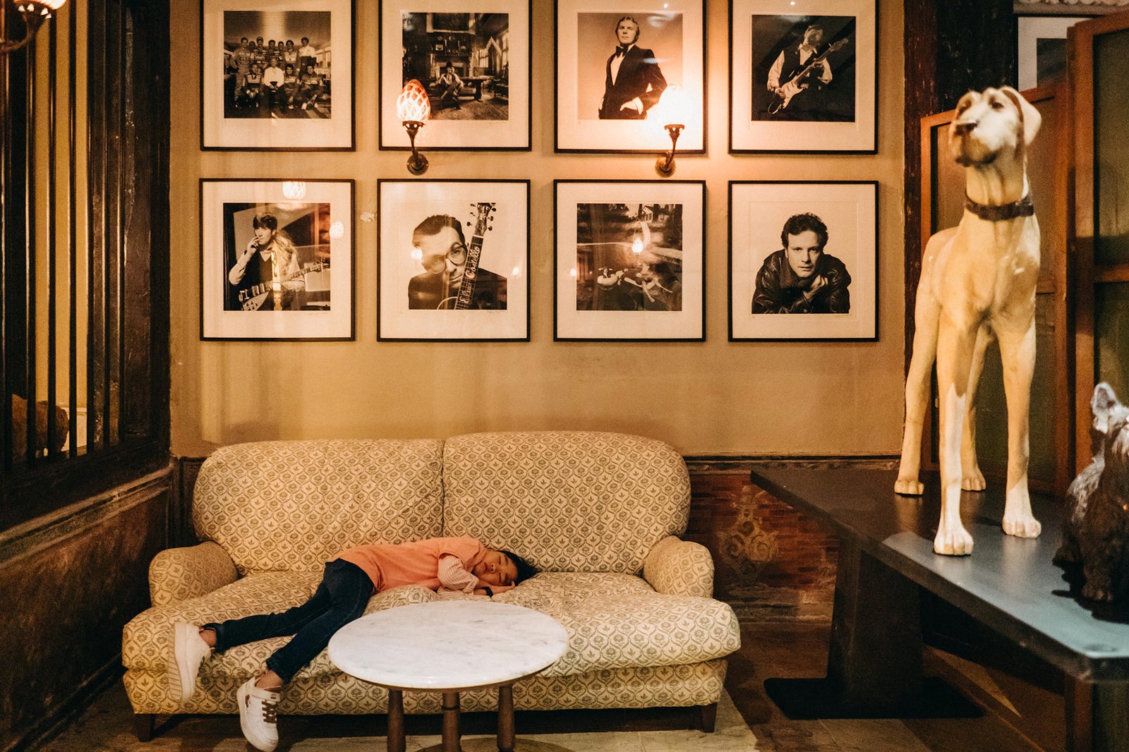 A young child sleeps on a patterned sofa in a dimly lit room, with framed black and white photos of musicians on the wall above. A tall dog statue stands to the right.