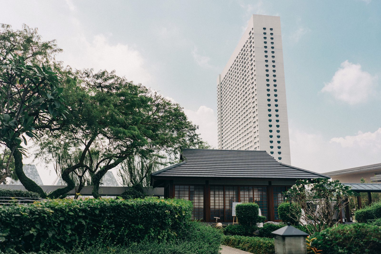 A serene Japanese garden pavilion with a dark tiled roof, surrounded by lush greenery and trees, stands in front of a tall, modern white skyscraper. This tranquil scene evokes the unique ambiance of a Keyaki Garden Pavilion wedding in Singapore.