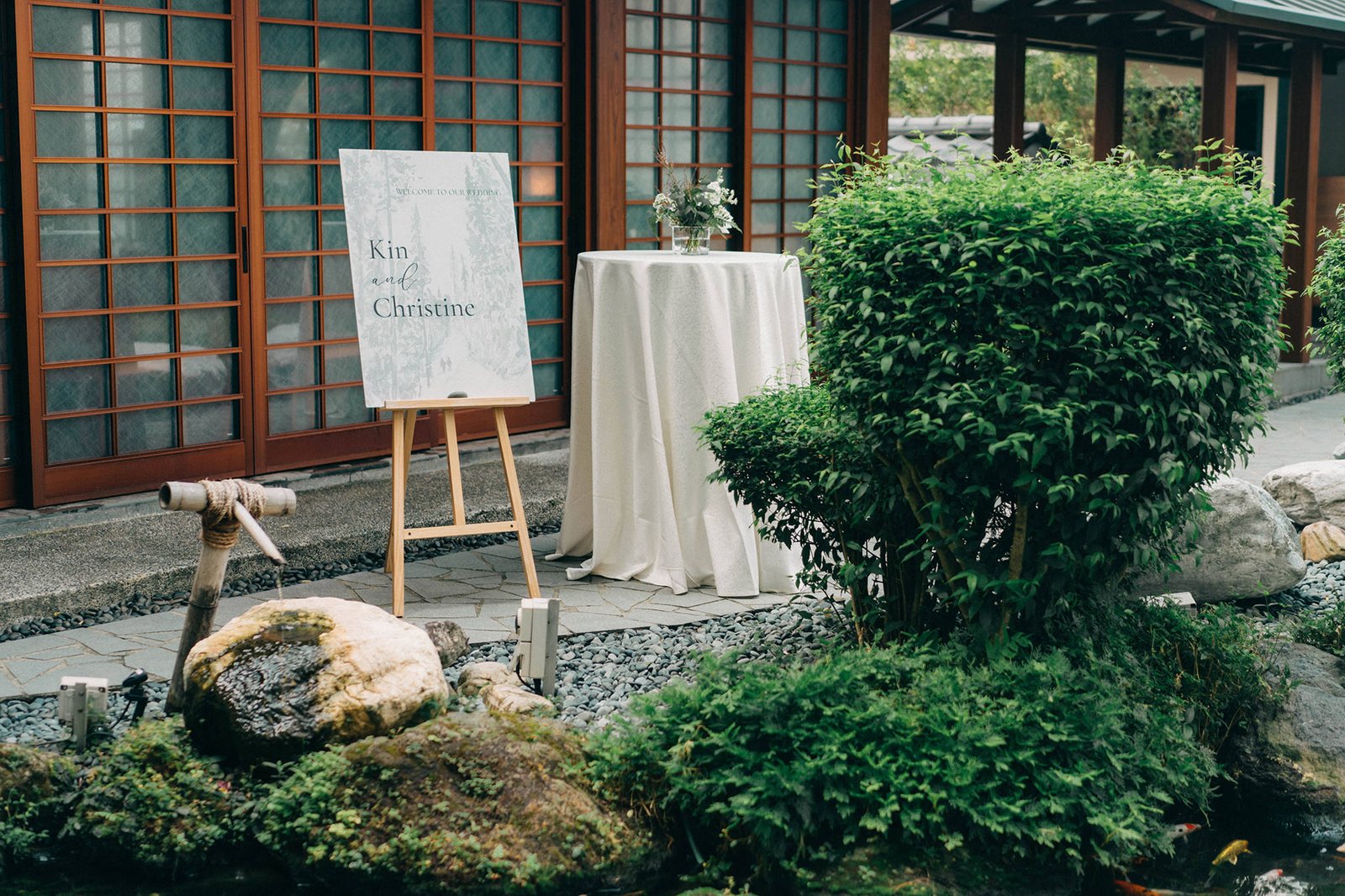 A serene wedding welcome sign for Kin and Christine stands on an easel in front of a Japanese-inspired Keyaki Garden Pavilion in Singapore, adorned with lush greenery and a round table setting.