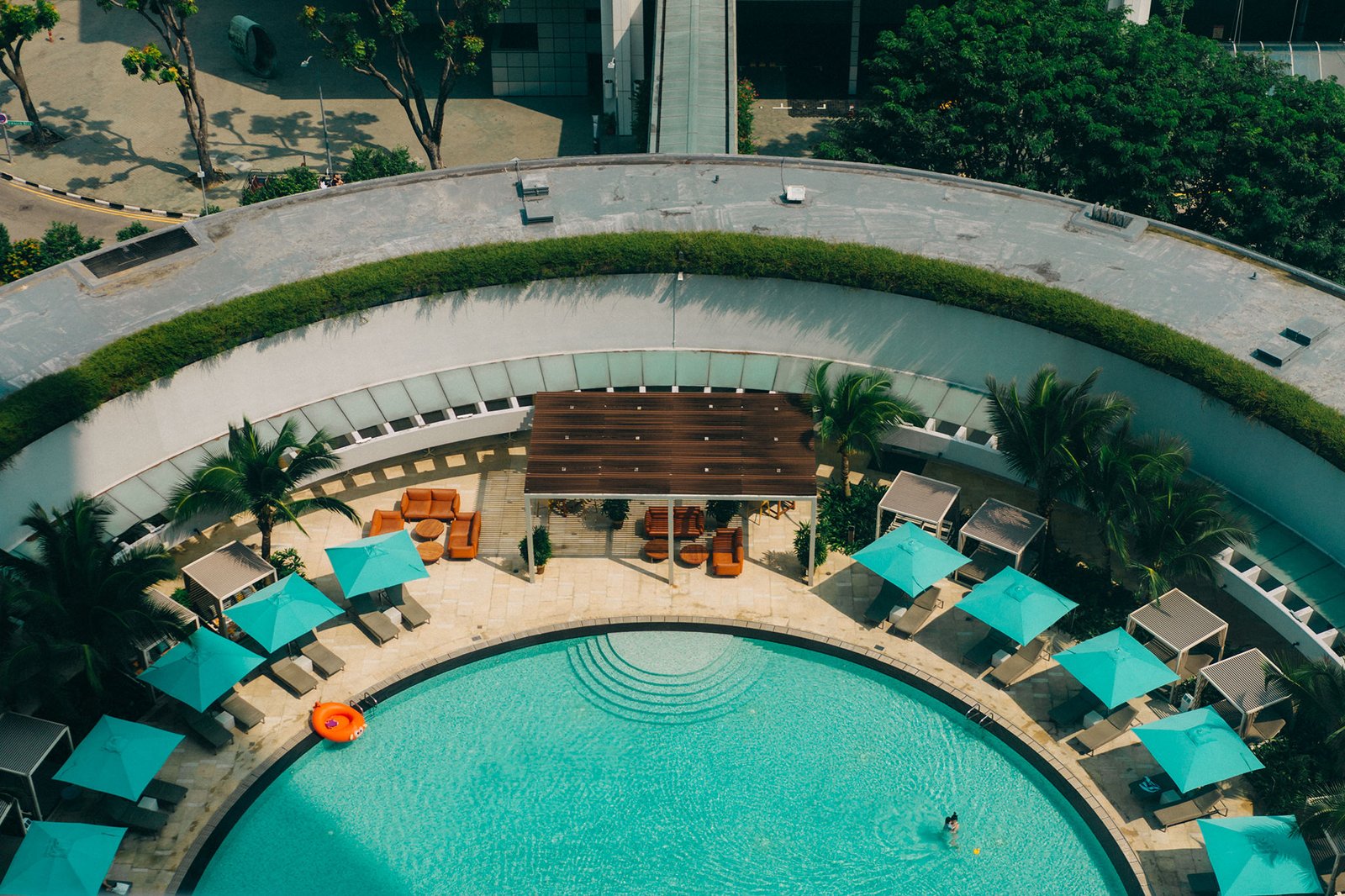 Aerial view of a luxurious outdoor swimming pool with turquoise water, surrounded by lush greenery and modern lounge areas with teal umbrellas. This serene setting could be ideal for events like a Keyaki Garden Pavilion Wedding in Singapore.