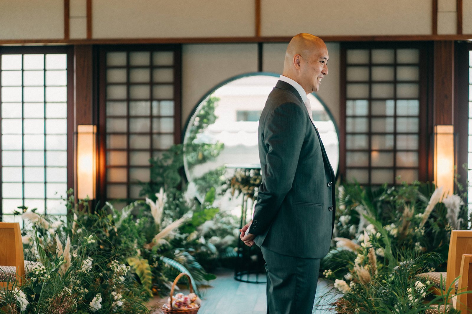 A groom in a dark suit smiles, hands clasped behind his back, at a Keyaki Garden Pavilion wedding in Singapore, surrounded by lush greenery and floral arrangements.