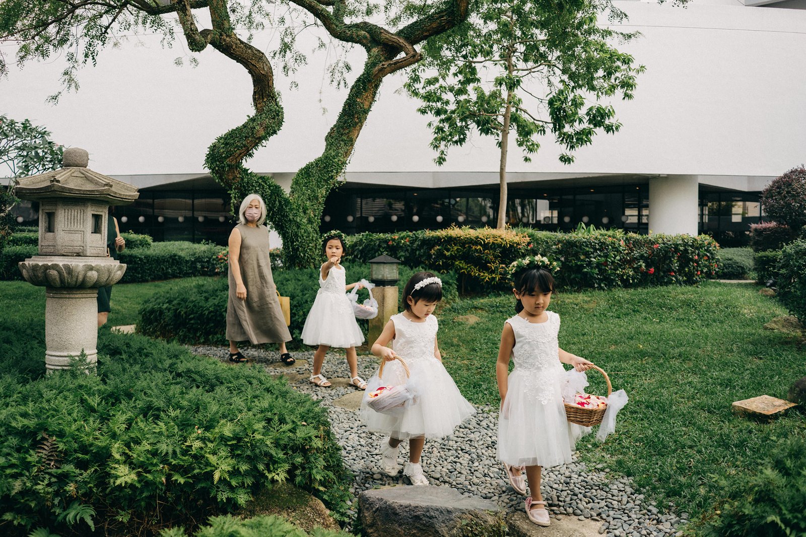 Flower girls in white dresses walk with baskets in a garden at a Keyaki Garden Pavilion wedding in Singapore, led by an adult.