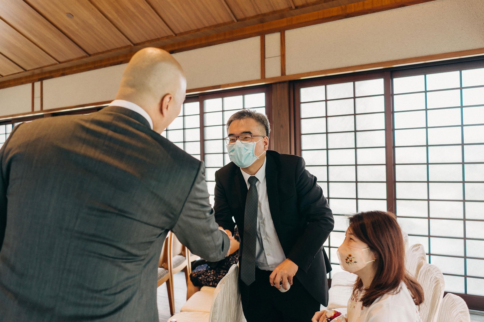 A man in a suit and mask shakes hands with another man in a suit and mask at a wedding reception, with a woman in a mask seated nearby. This scene is from the Keyaki Garden Pavilion Wedding Singapore.