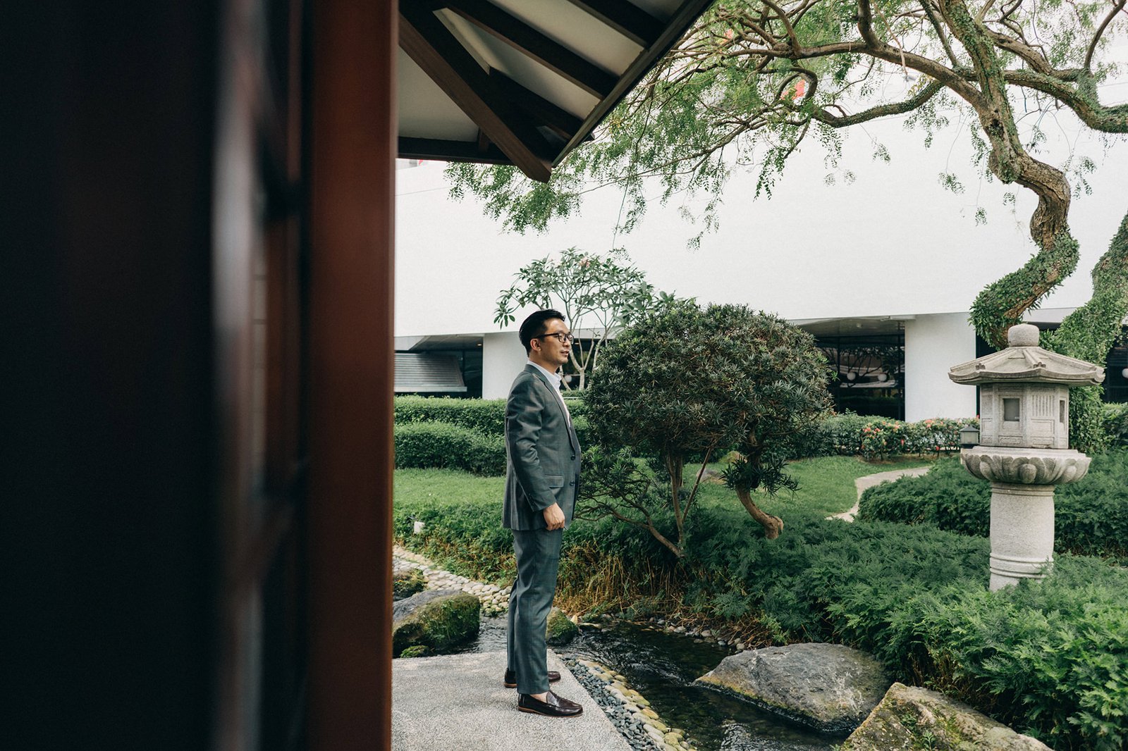 Groom in a grey suit stands outside the Keyaki Garden Pavilion in Singapore, looking to the side with lush greenery and a stone lantern in the background.