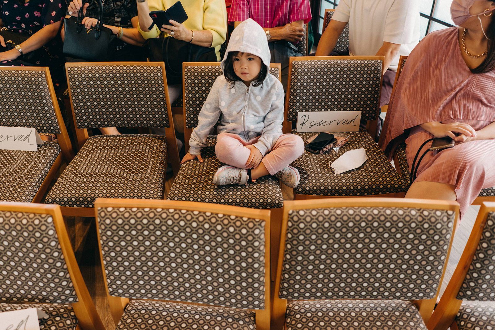A young girl in a grey hoodie sits cross-legged on a reserved chair at the Keyaki Garden Pavilion wedding in Singapore, looking towards the camera.