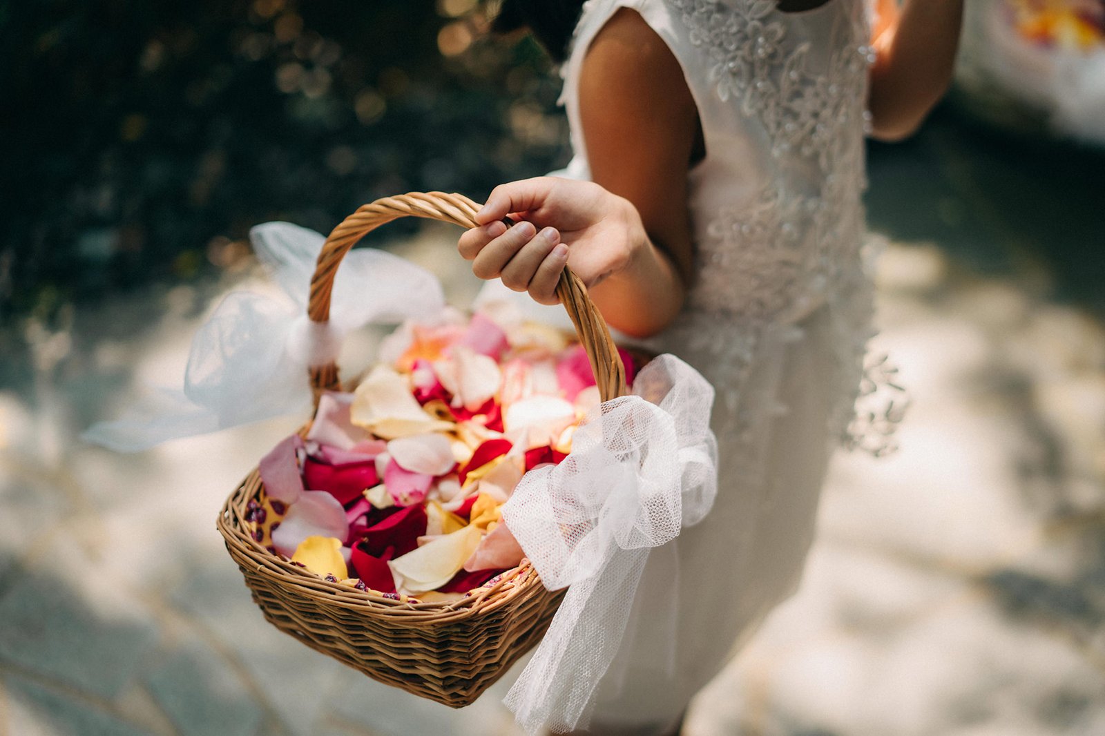 A flower girl in a white dress holds a woven basket filled with colorful rose petals, ready to scatter them at a Keyaki Garden Pavilion wedding in Singapore.