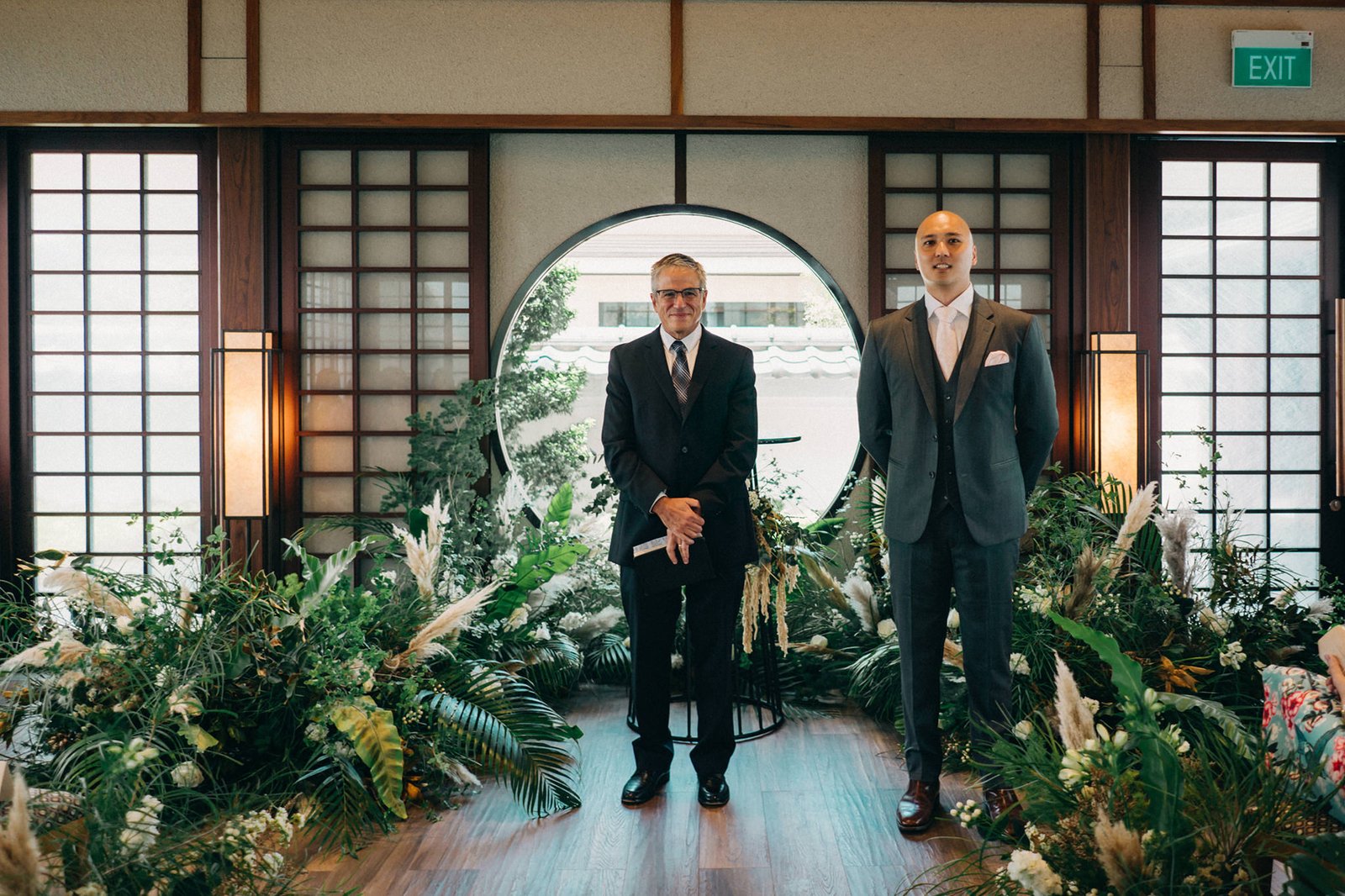 Two men in formal wedding attire stand in a lush, green ceremony setting at a Keyaki Garden Pavilion Wedding in Singapore, with a circular window and Japanese-inspired architecture in the background.