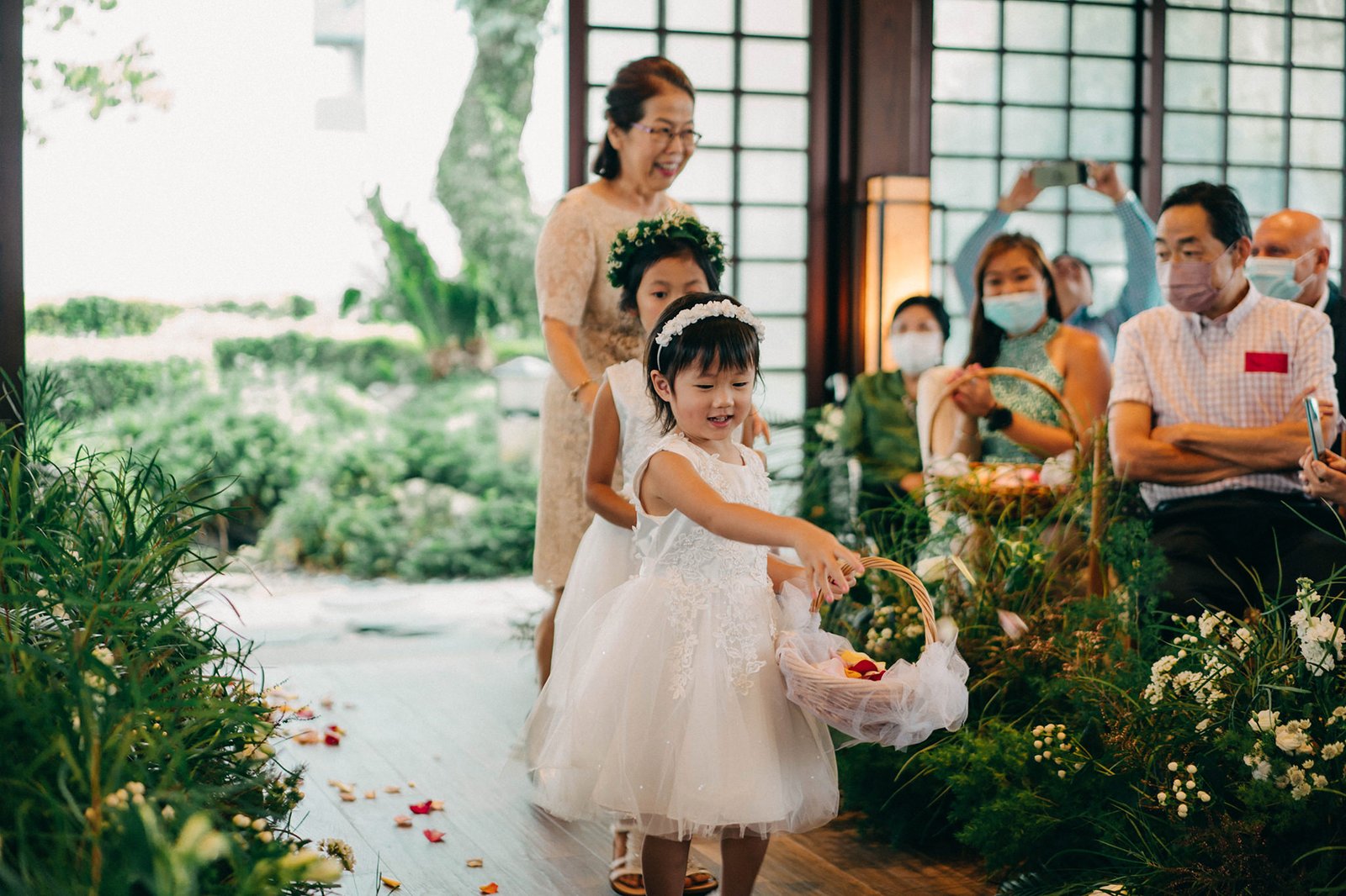 Flower girls in white dresses walk down the aisle at a Keyaki Garden Pavilion wedding in Singapore, scattering petals from baskets. A woman in a beige dress and guests watch.