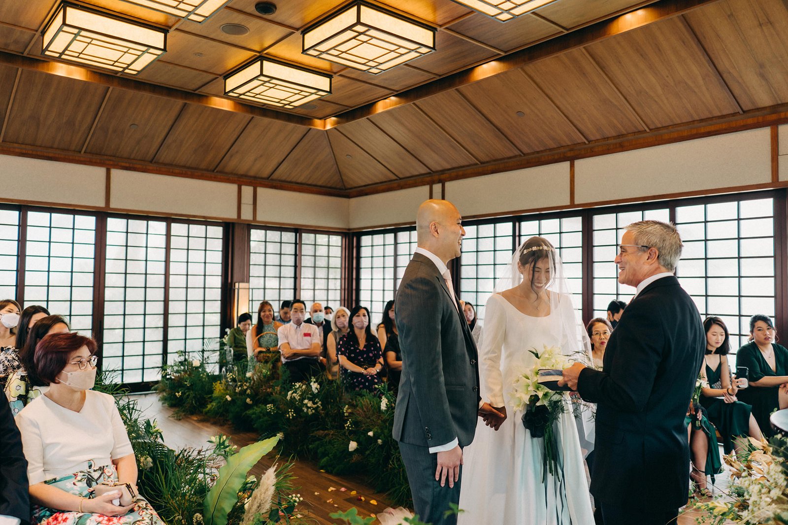 A wedding ceremony at the Keyaki Garden Pavilion in Singapore, with a bride and groom exchanging vows while officiant looks on. Guests are seated in a lush, green setting.