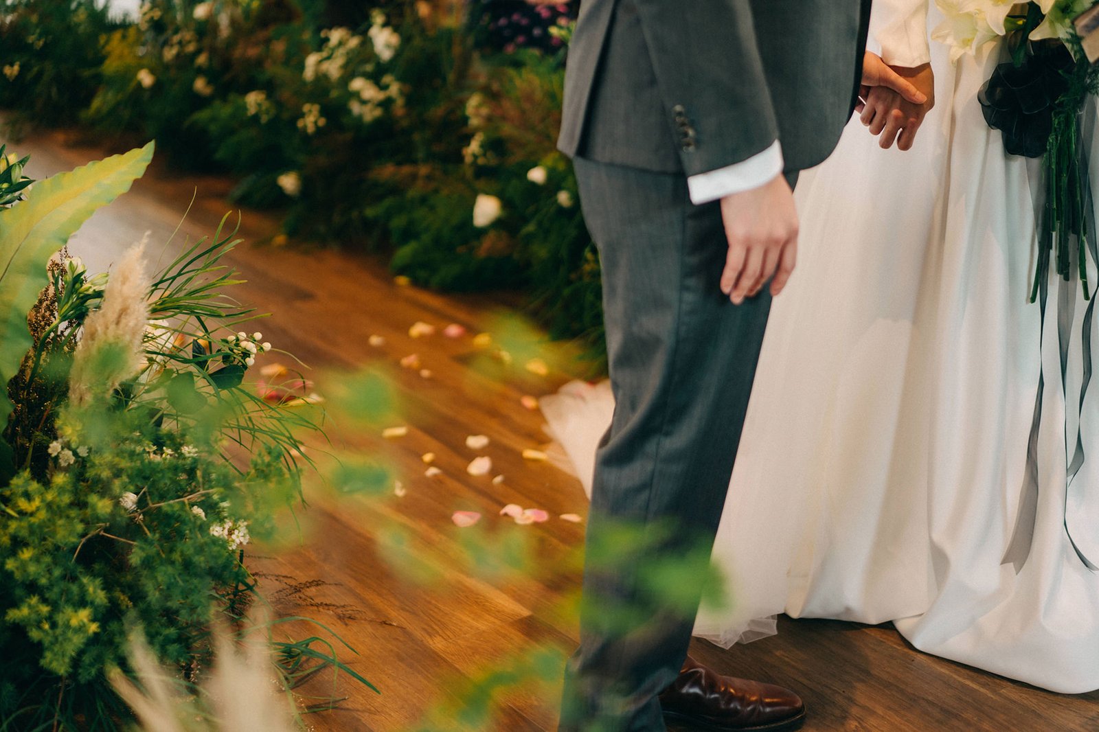 A bride in a white gown and groom in a grey suit hold hands during a wedding ceremony at the Keyaki Garden Pavilion in Singapore, with lush greenery and fallen petals.
