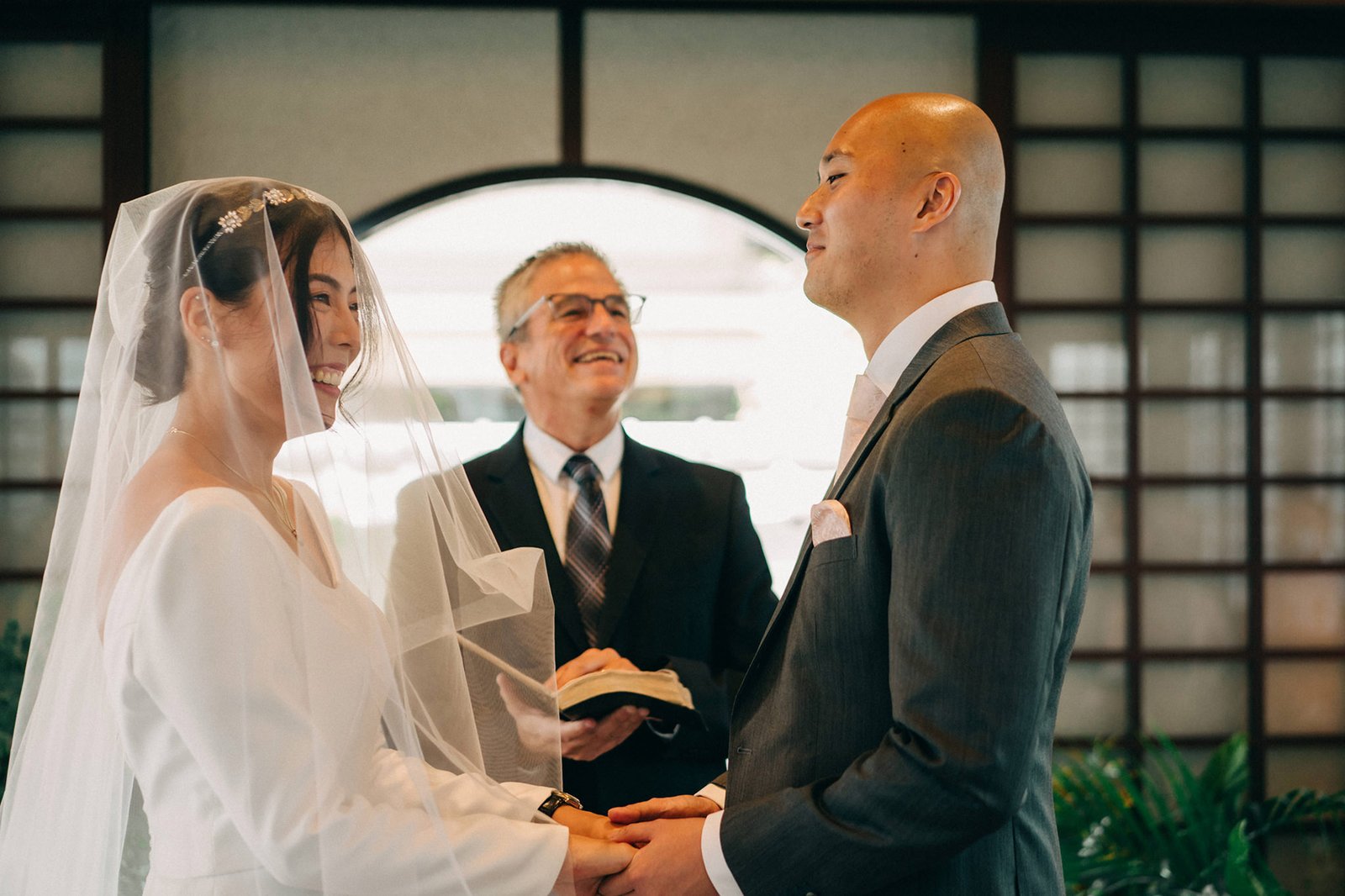 A smiling bride in a white gown and veil, and a groom in a grey suit, hold hands during their wedding ceremony at the Keyaki Garden Pavilion in Singapore, with an officiant in the background.