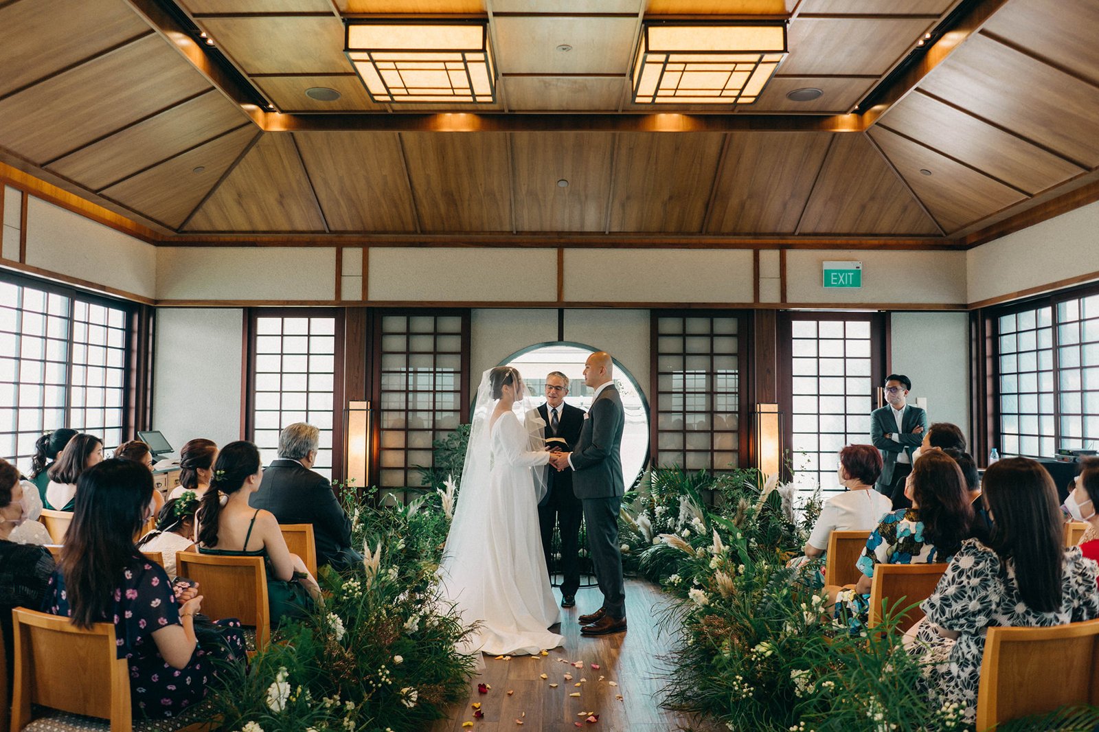 Kin + Christine's intimate wedding ceremony at Keyaki Garden Pavilion, Singapore. The couple exchanges vows under a circular arch, surrounded by lush greenery and seated guests.