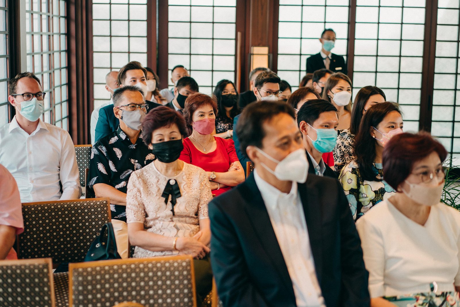 Guests in masks attend a wedding ceremony at the Keyaki Garden Pavilion in Singapore. People in formal attire are seated, facing forward, with large windows in the background.