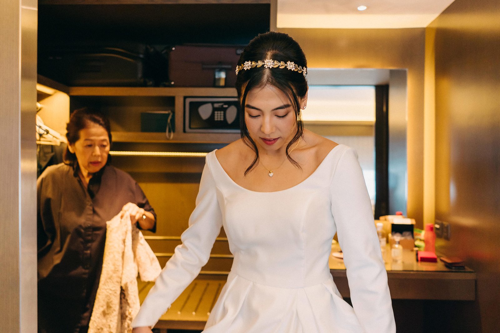 A bride in a white wedding dress and tiara looks down, while an older woman in the background holds a lace garment. This intimate moment captures pre-wedding preparations.