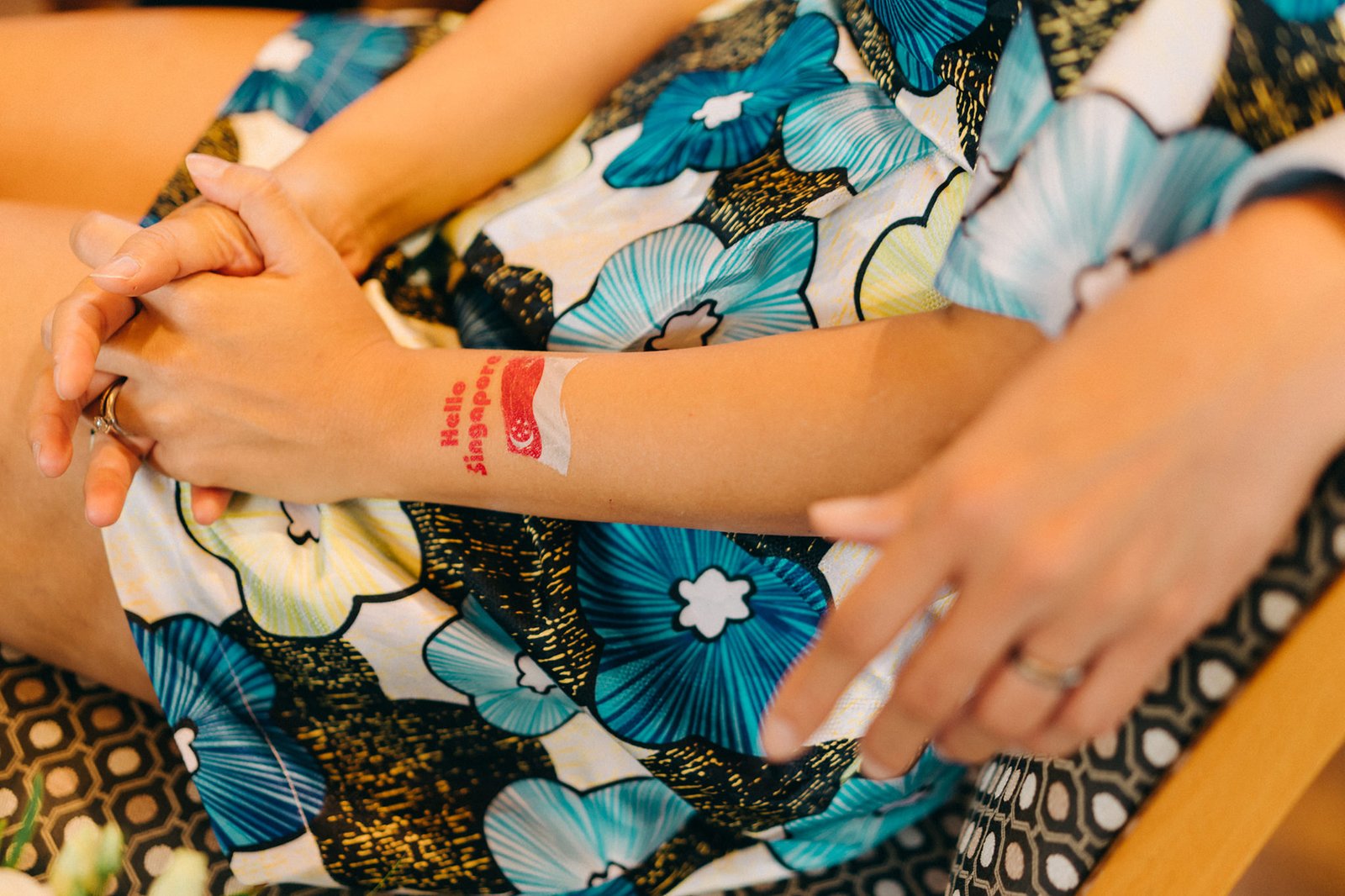 Close-up of a bride's arm with a "Hello Singapore" temporary tattoo, resting on her lap in a floral dress at a Keyaki Garden Pavilion wedding in Singapore.