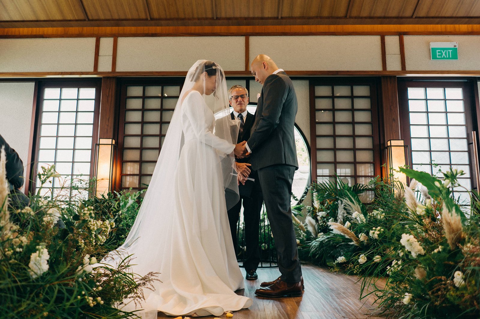 A wedding ceremony at Keyaki Garden Pavilion Singapore, with a bride in a white gown and veil holding hands with a groom in a suit, officiated by a celebrant amidst lush greenery.