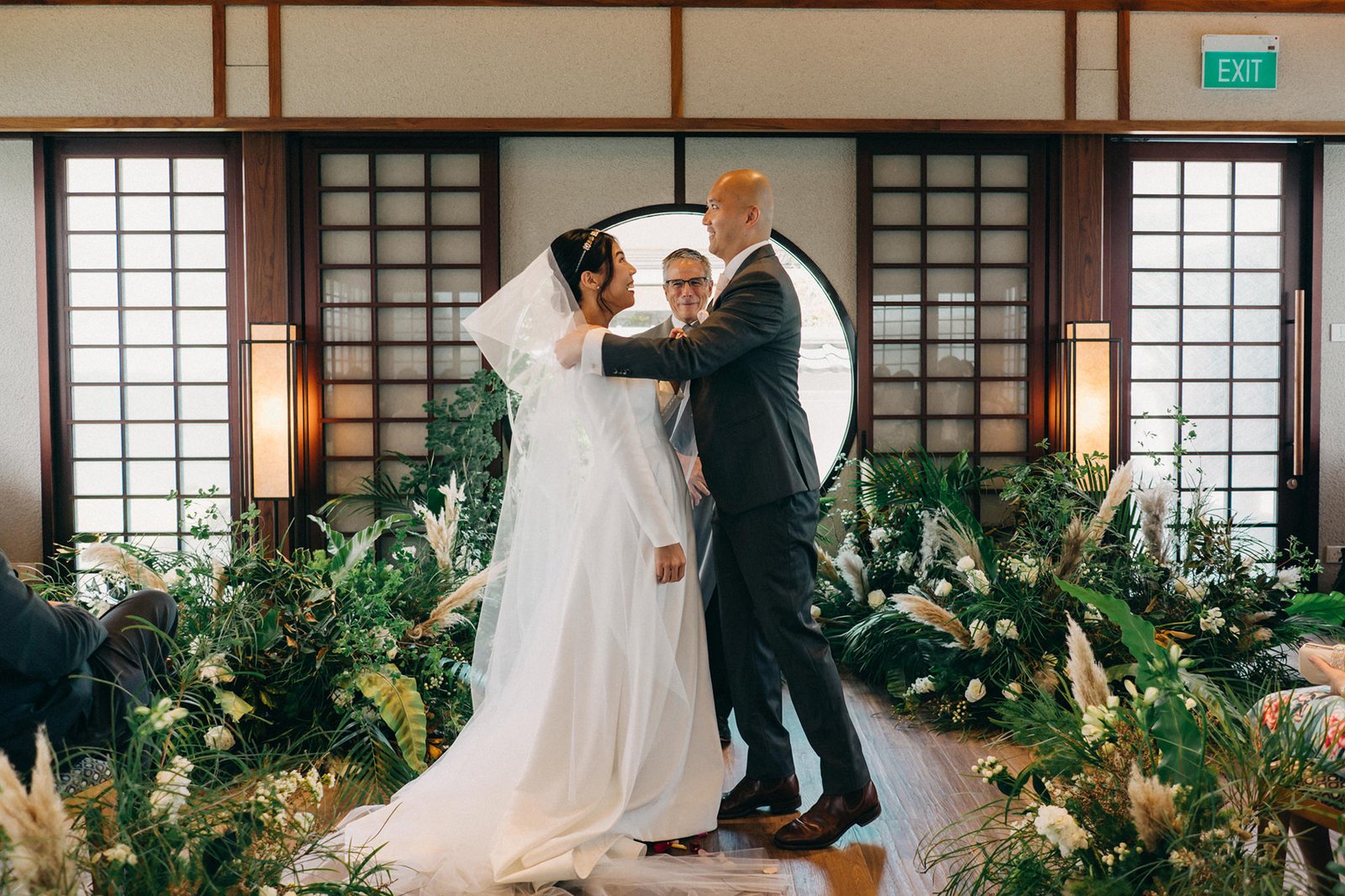 A joyful wedding ceremony at Keyaki Garden Pavilion, Singapore, as a bride in a white gown and veil exchanges vows with her groom, officiated by a celebrant. Lush greenery and pampas grass surround the couple.