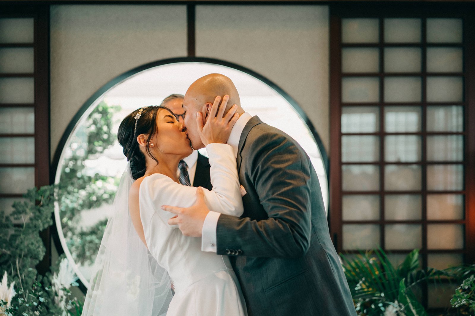 A bride and groom share a tender kiss at their Keyaki Garden Pavilion wedding in Singapore, with lush greenery and a circular window framing the intimate moment.