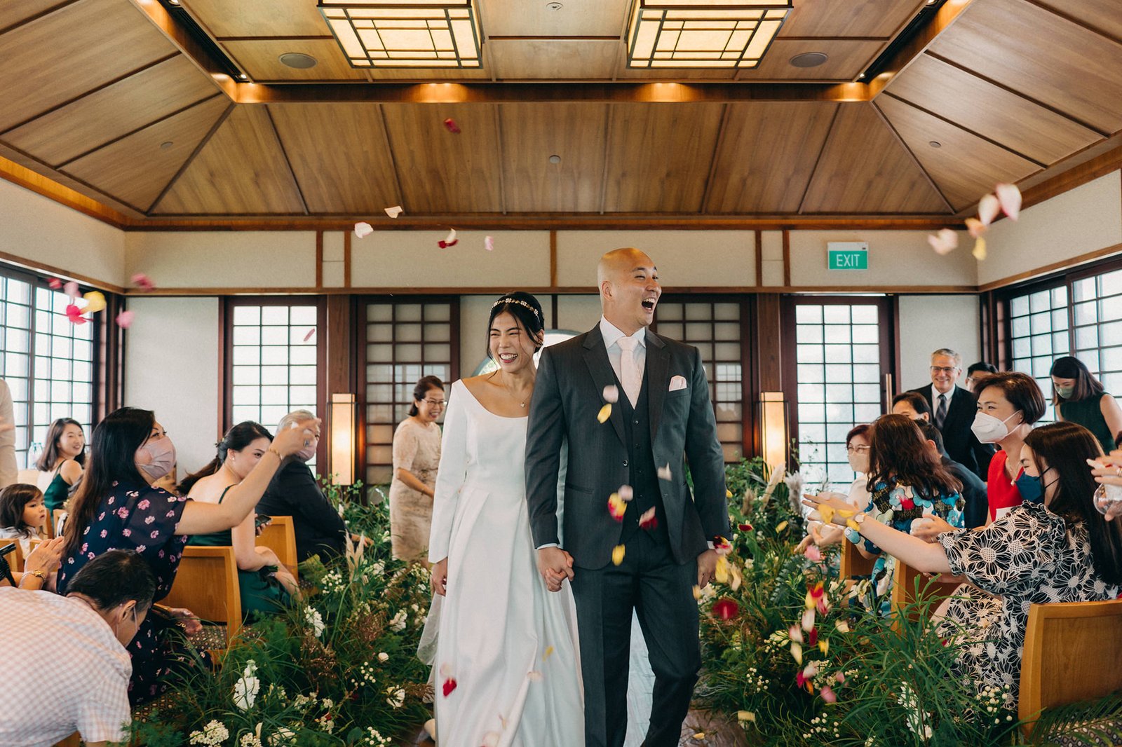 A joyous bride and groom walk down an aisle showered with flower petals at their Keyaki Garden Pavilion wedding in Singapore, surrounded by smiling guests.