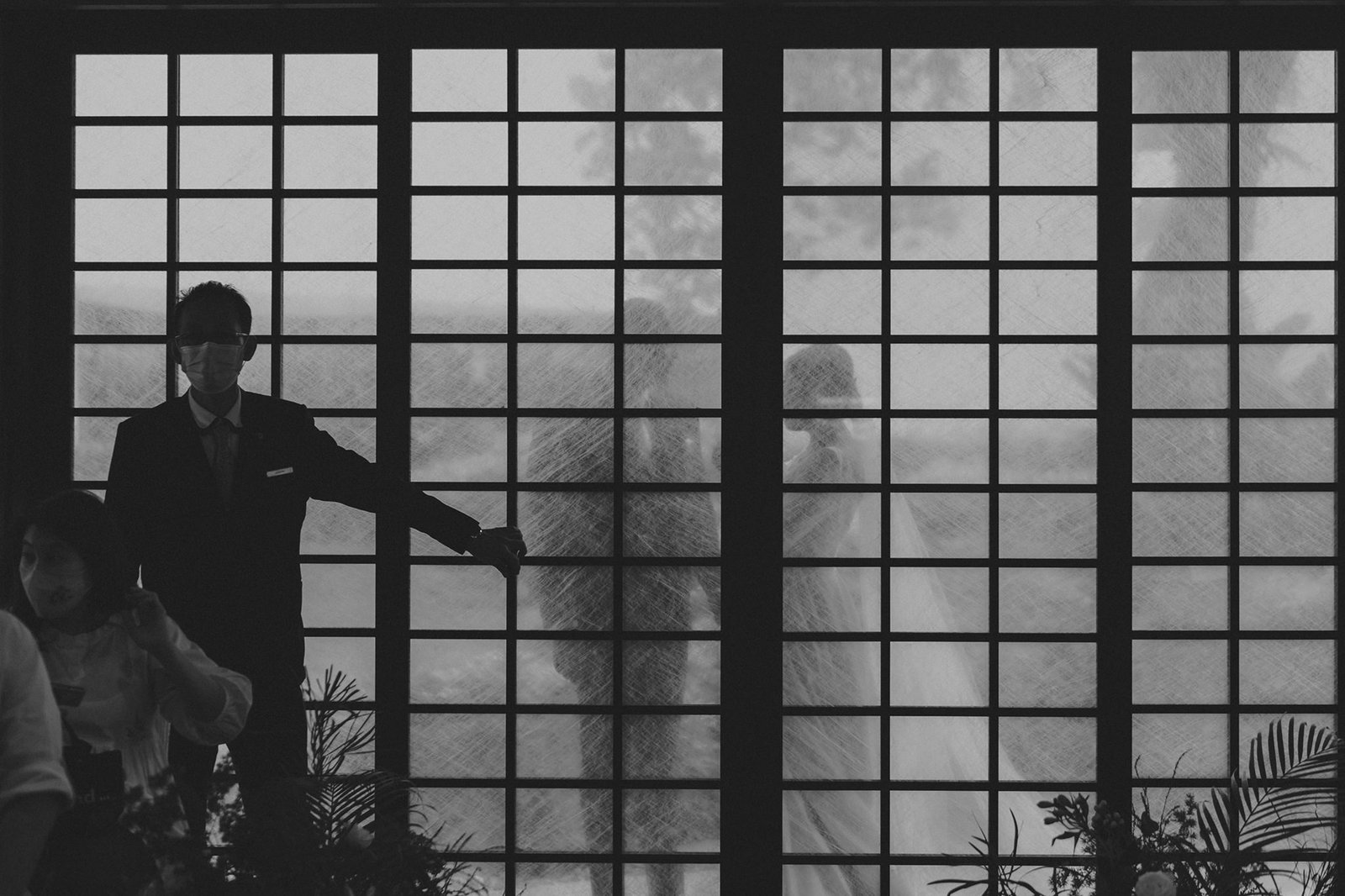 Groom in tuxedo and mask stands near a window at Keyaki Garden Pavilion Wedding Singapore, with bride in veil blurred in background.