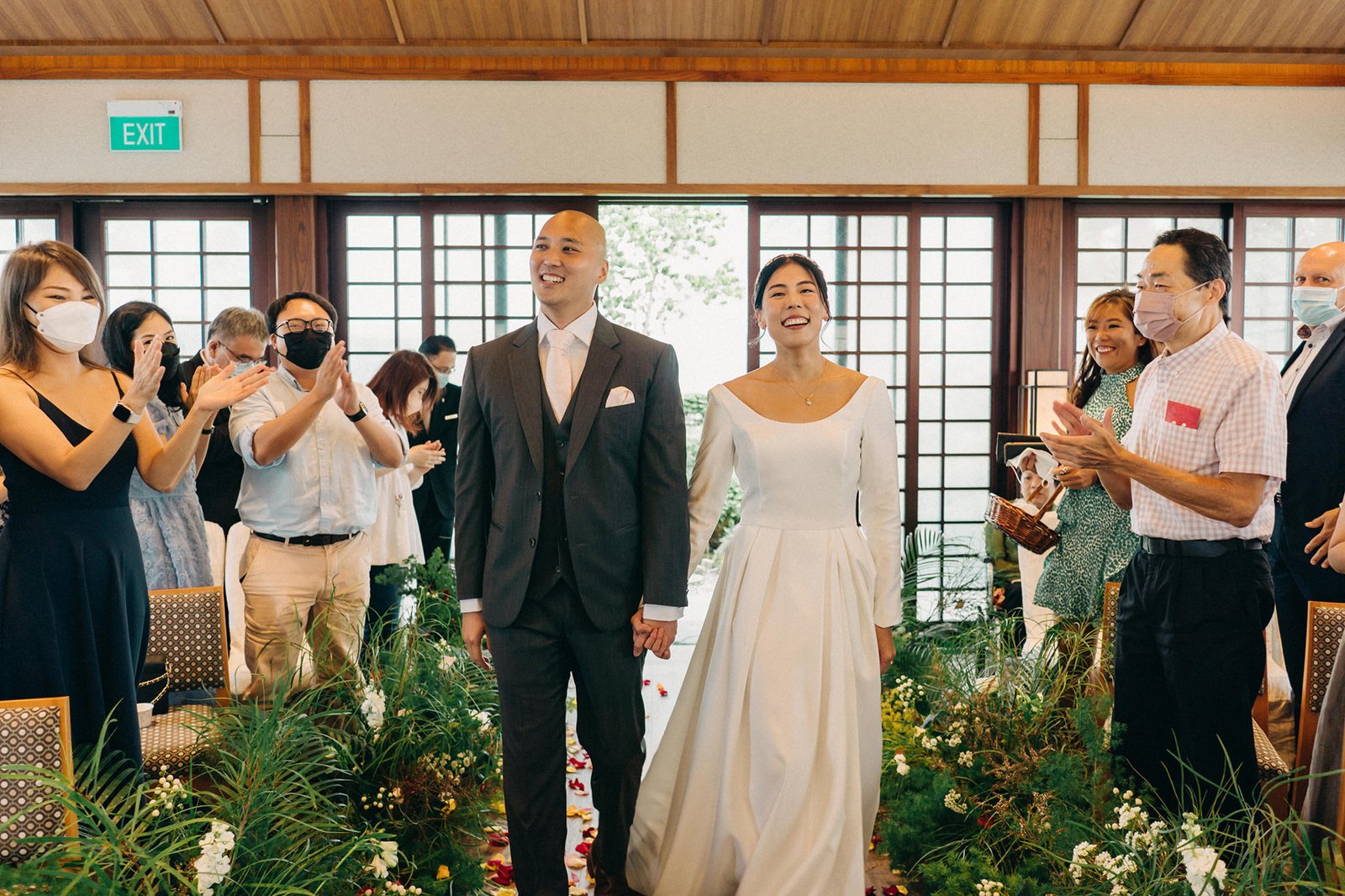 A happy bride in a white dress and groom in a suit walk down the aisle at a Keyaki Garden Pavilion wedding in Singapore, holding hands as guests watch.