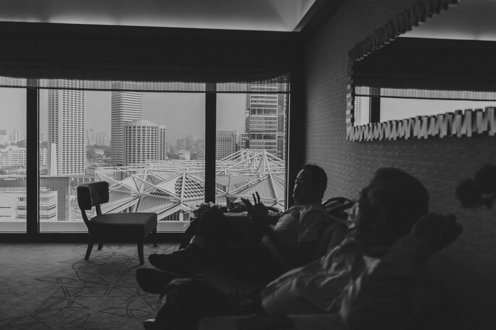 Two men relax in a dimly lit hotel room with a panoramic city view, hinting at a Keyaki Garden Pavilion wedding in Singapore.