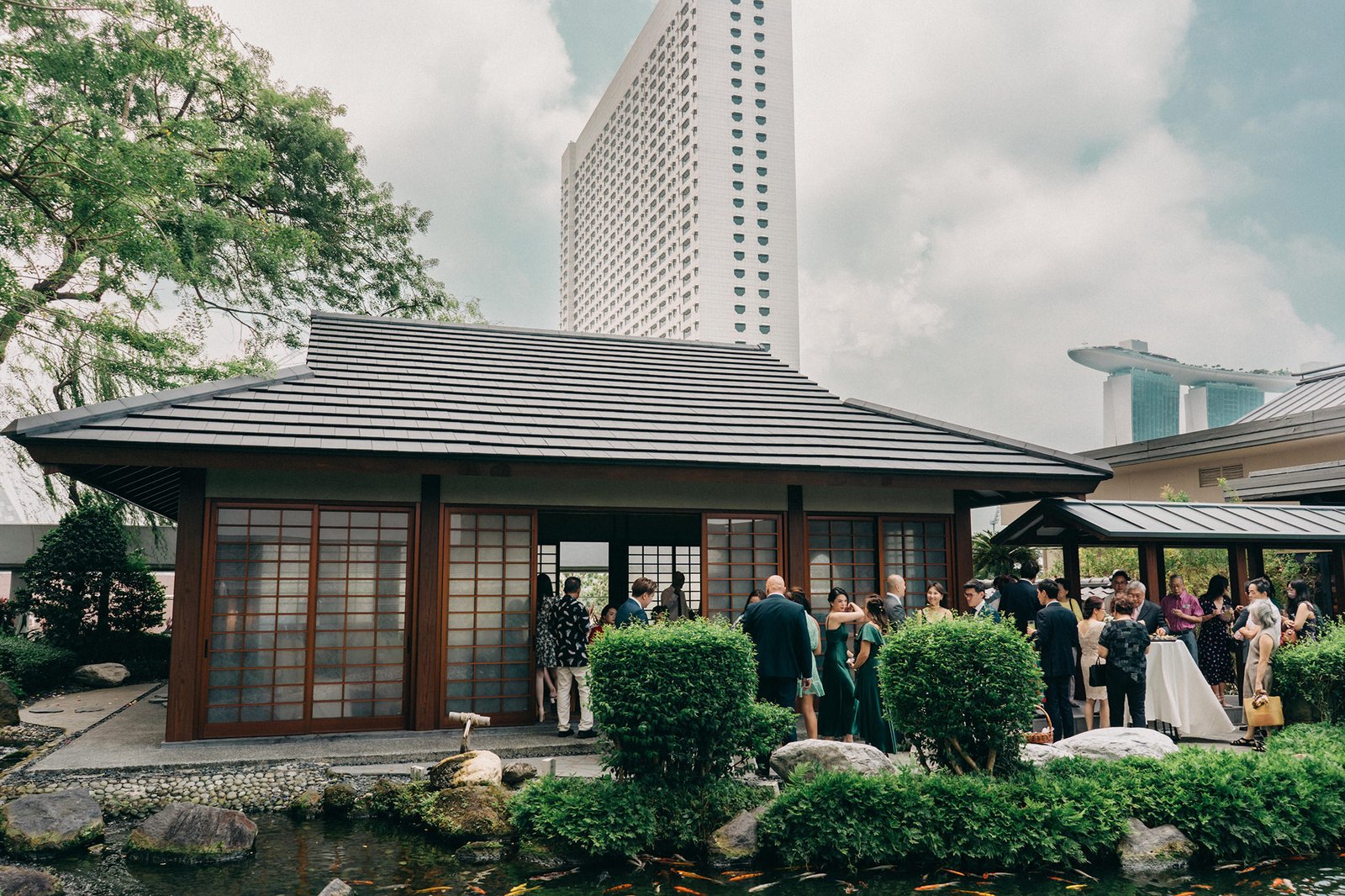 A wedding reception at the Keyaki Garden Pavilion in Singapore, with guests mingling near a traditional Japanese-style building and koi pond, modern skyscrapers in the background.