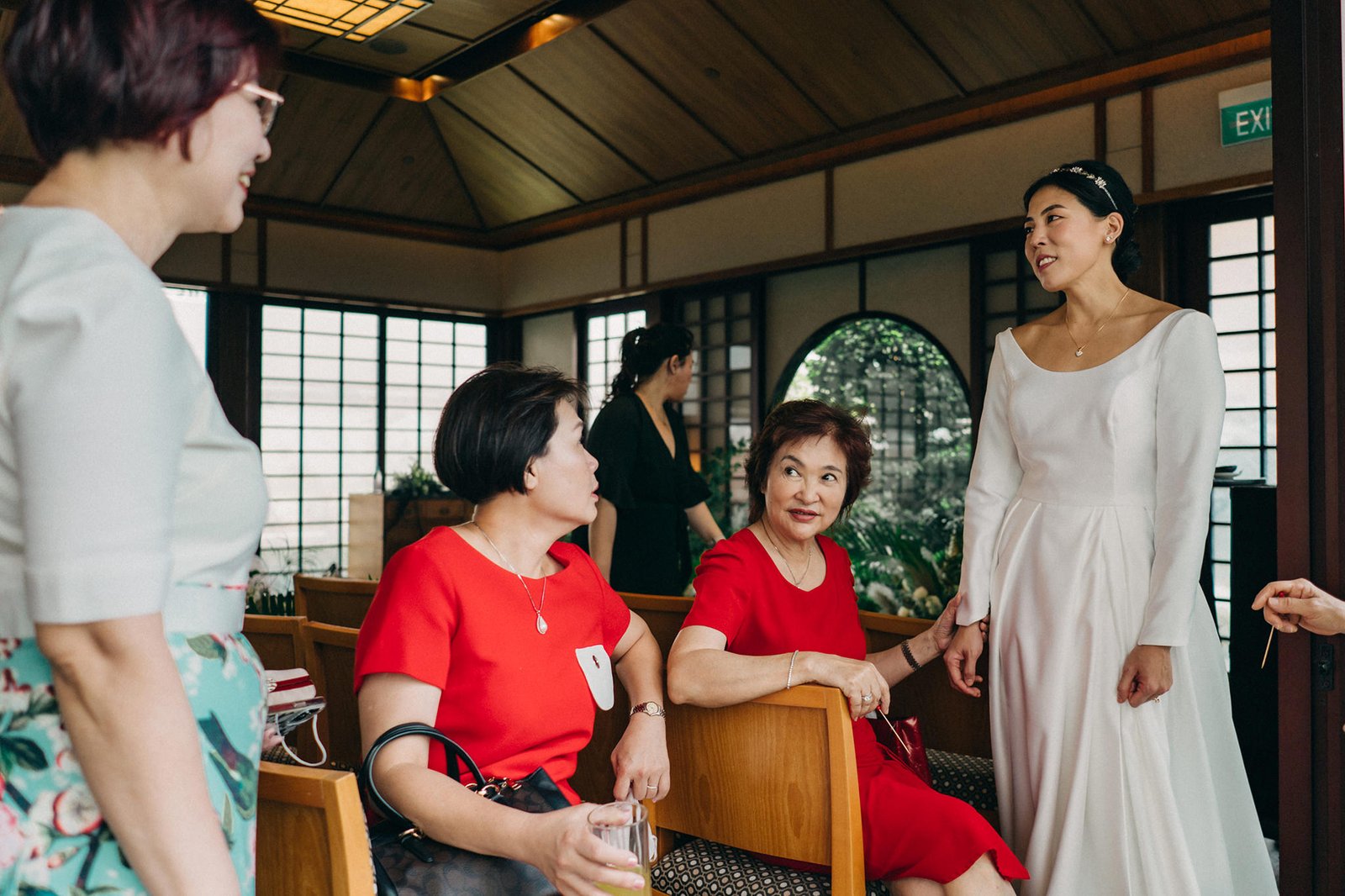Bride in a white gown smiles at guests at a Keyaki Garden Pavilion wedding in Singapore. Two women in red and a woman in white converse in a warm, wood-paneled room.