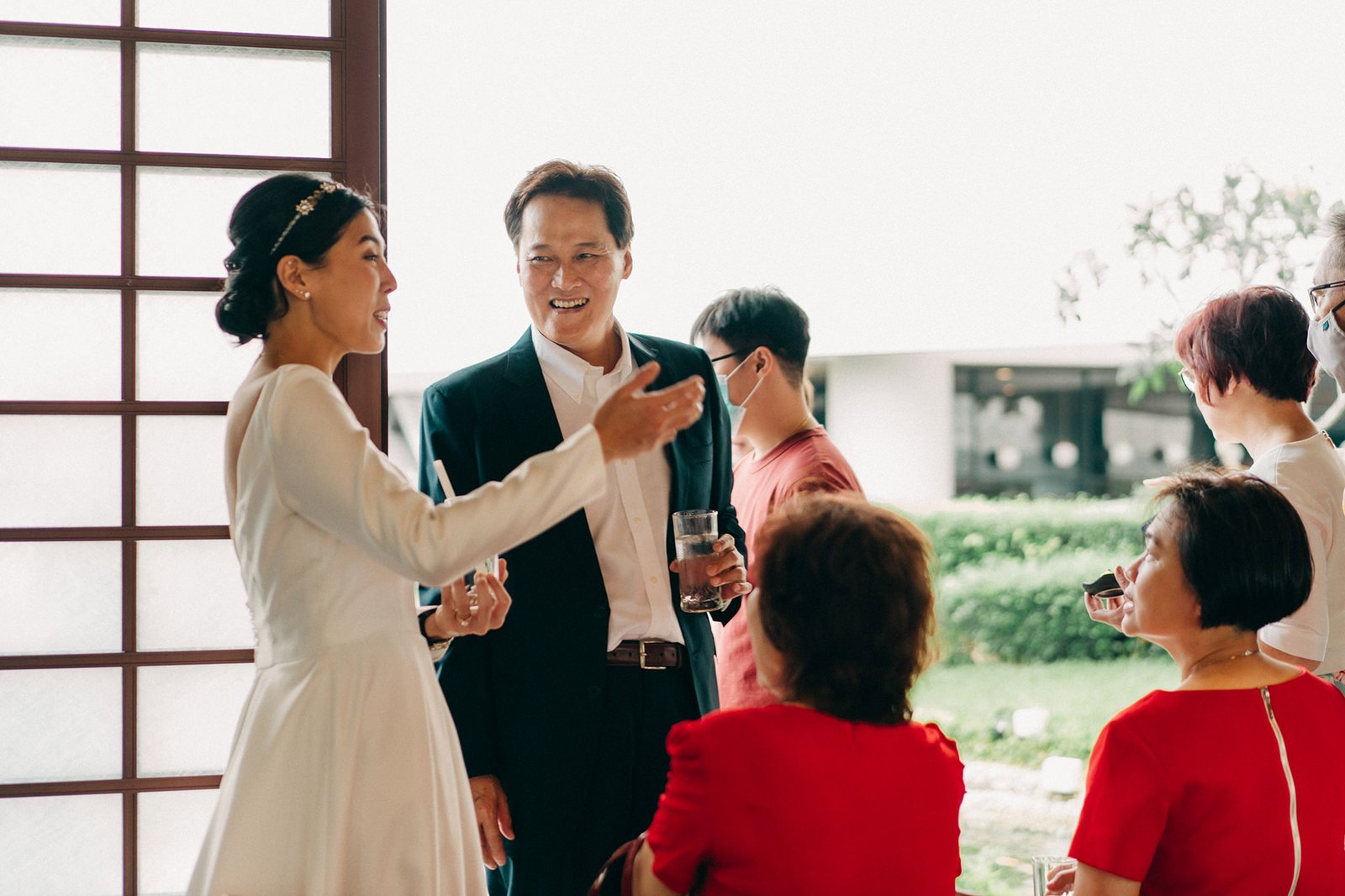 A bride in a white dress and a man in a suit converse warmly with guests at a wedding reception at the Keyaki Garden Pavilion, Singapore.