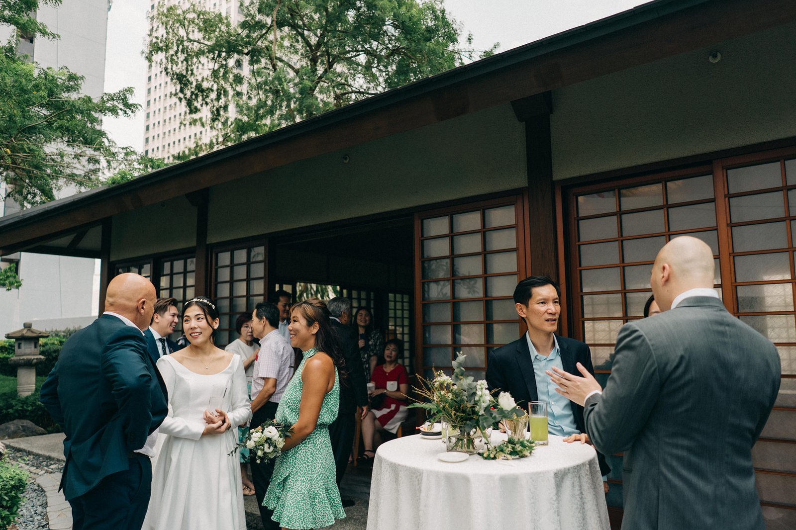 Kin and Christine's intimate wedding reception at Keyaki Garden Pavilion, Singapore. Guests mingle near a traditional Japanese-style building, with a bride in a white dress and groom in a suit.