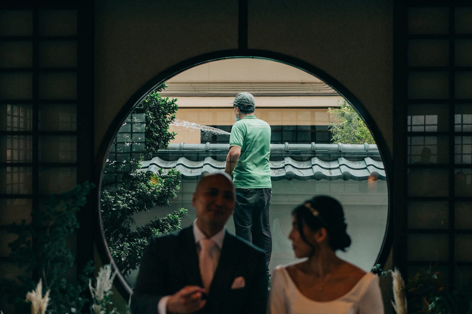 A wedding couple is blurred in the foreground, looking towards a man watering plants through a circular window at a Keyaki Garden Pavilion wedding in Singapore.