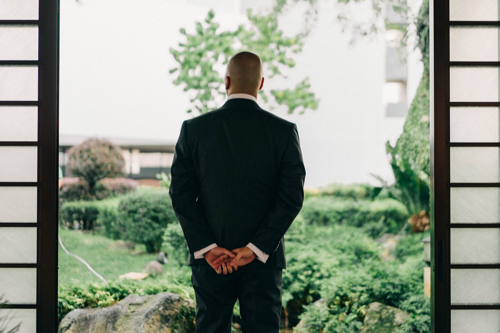 Groom in a dark suit stands with hands clasped behind his back, looking out at the lush greenery of the Keyaki Garden Pavilion in Singapore.