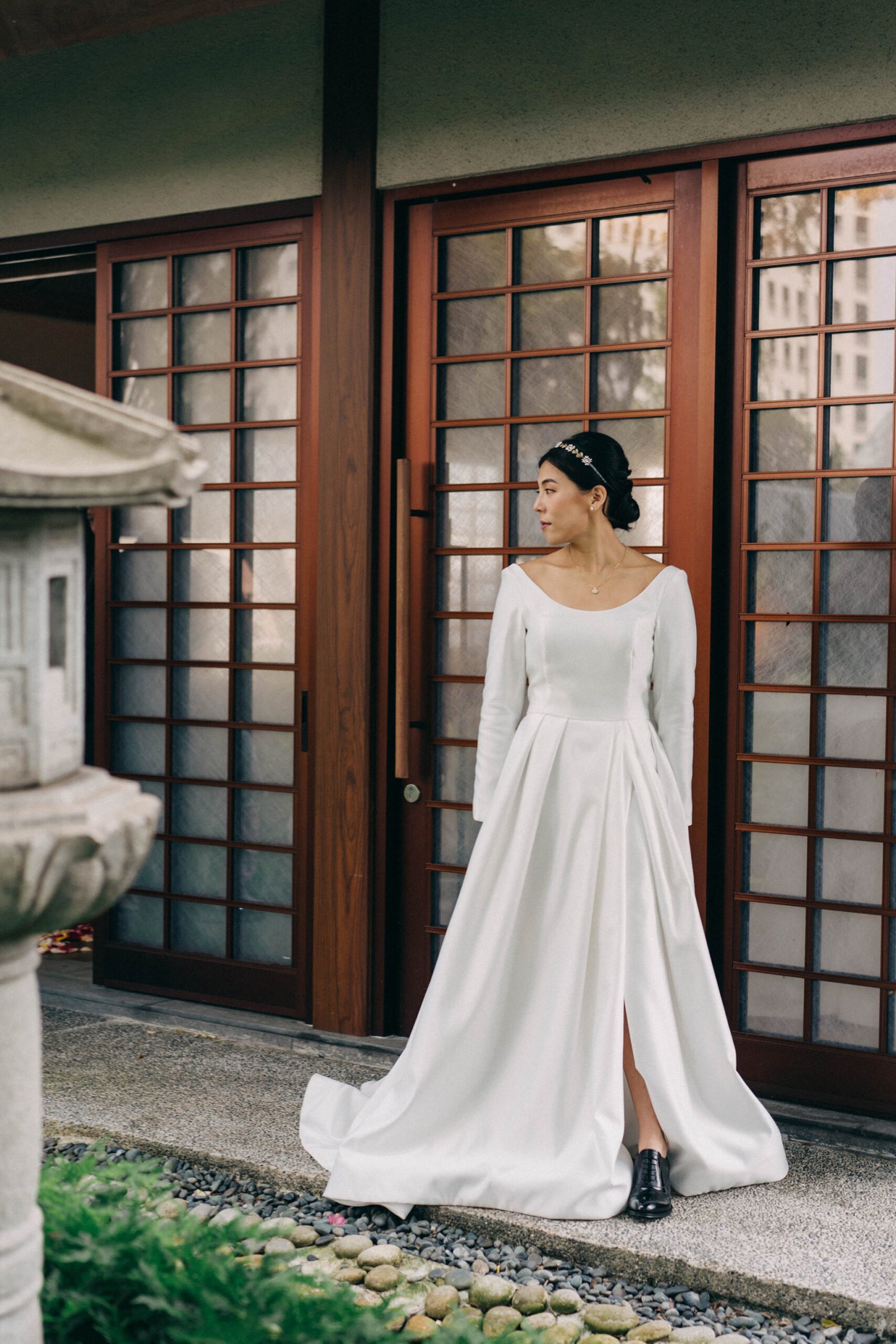 A bride in a white satin wedding dress with a high slit stands in front of traditional Japanese sliding doors at a Keyaki Garden Pavilion wedding in Singapore.