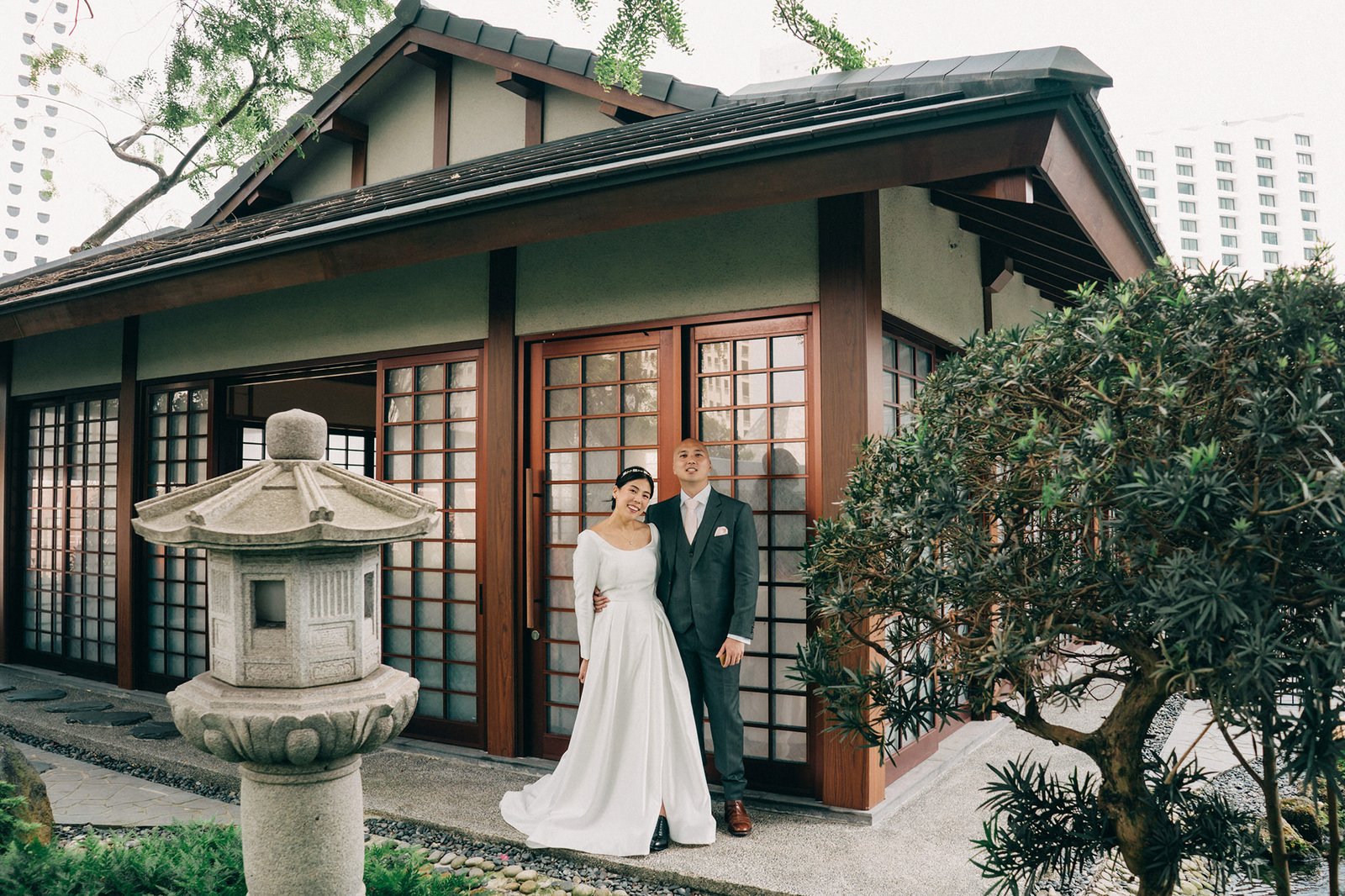 A happy couple, Kin and Christine, pose for their wedding portrait at the Keyaki Garden Pavilion in Singapore. The bride wears a long-sleeved white wedding dress, and the groom is in a dark suit with a pink tie, standing in front of a traditional Japanese-style building with sliding doors and a stone lantern.