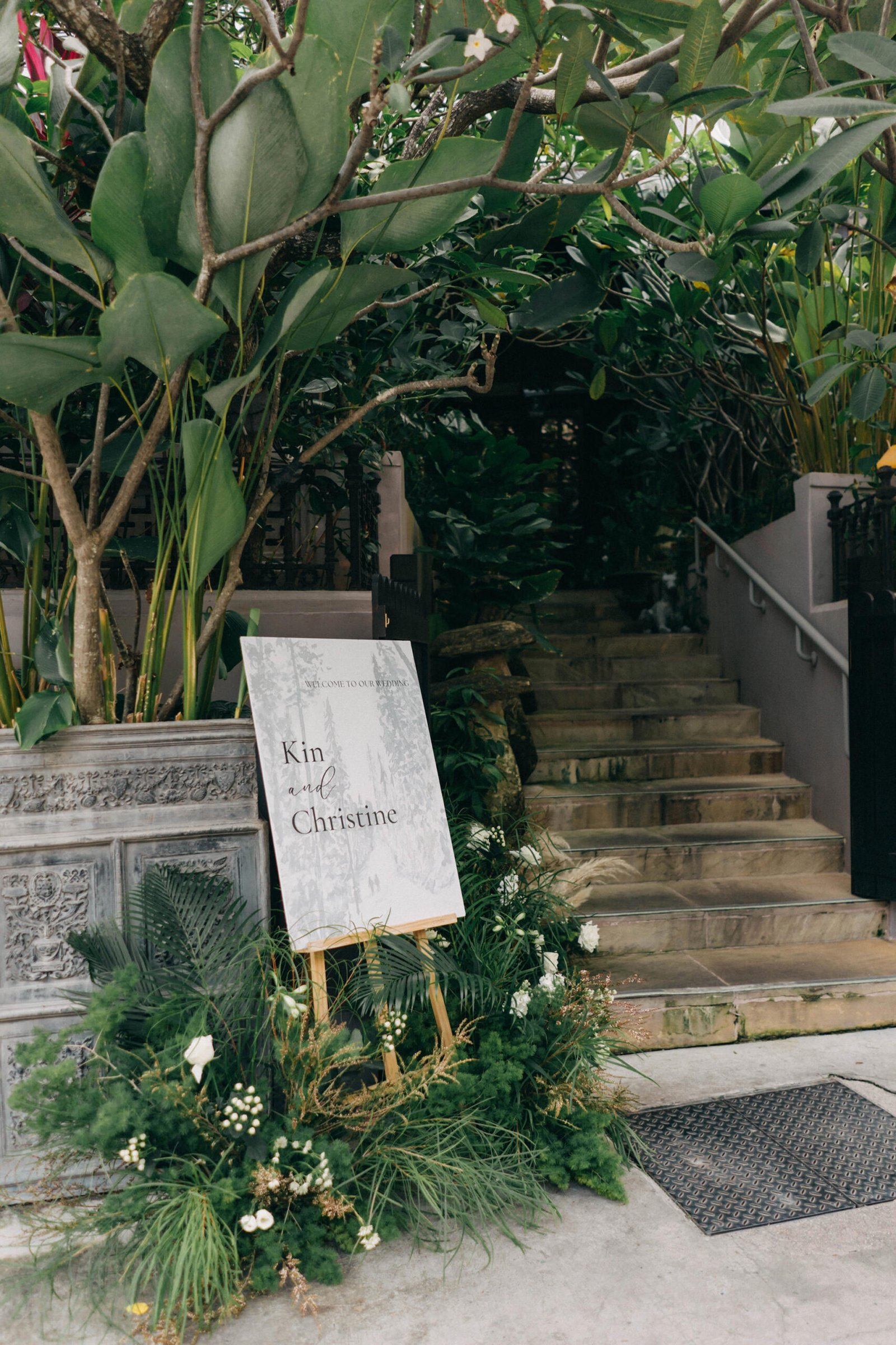 A wedding welcome sign for Kin and Christine stands at the entrance of The English House Wedding Singapore, adorned with lush greenery and white flowers, leading up a stone staircase.