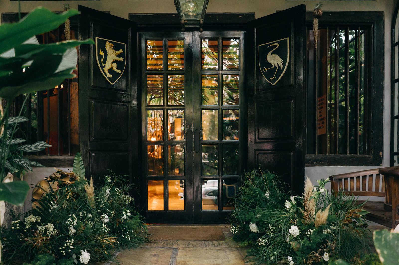Elegant black double doors with golden crests flank a sunlit entrance to The English House, a wedding venue in Singapore, adorned with lush greenery and white flowers.