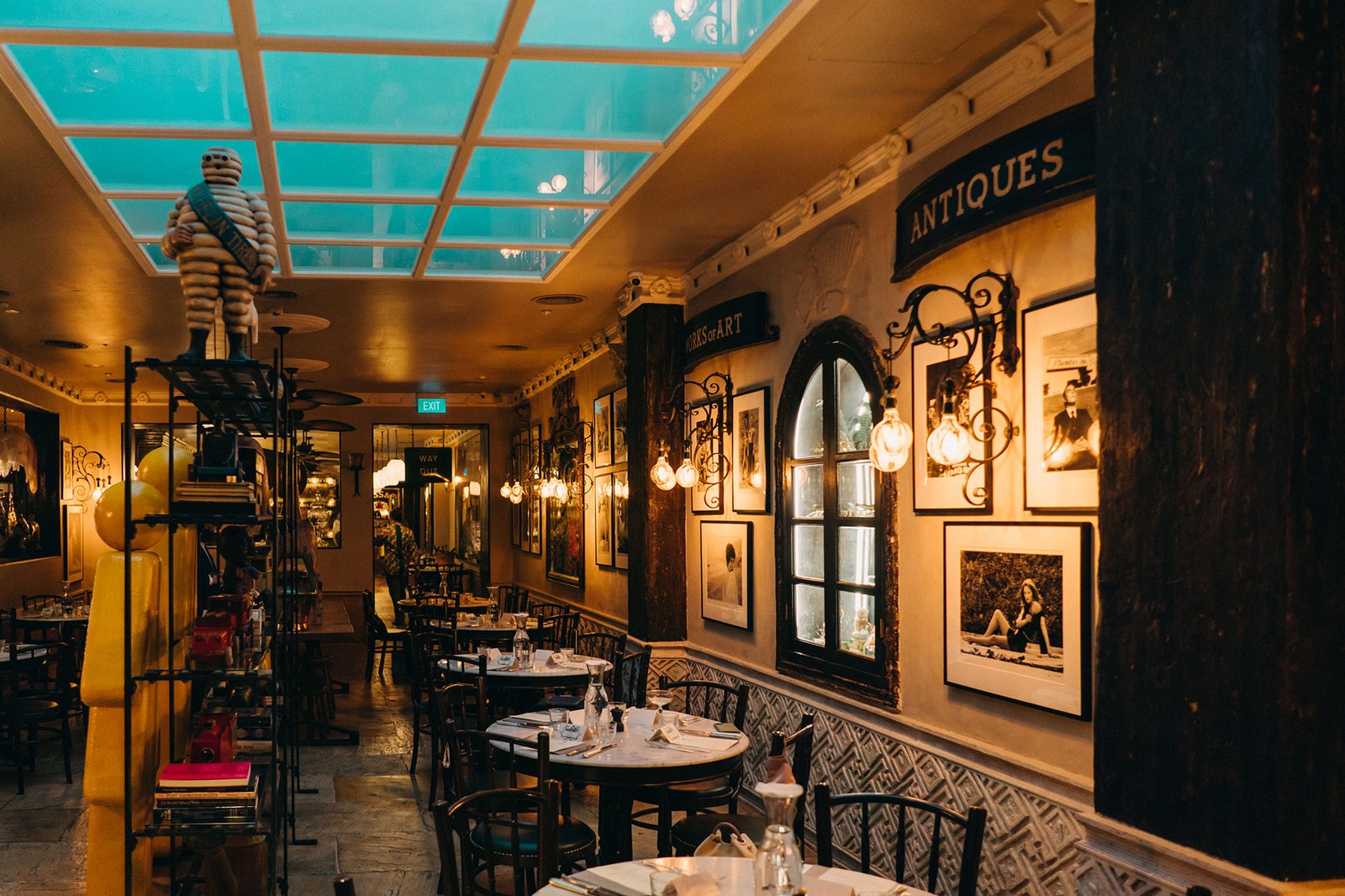 Interior of a dimly lit, elegant restaurant with a glass ceiling, featuring round tables set for dining, antique decor, and framed photographs on the walls. The ambiance evokes a sophisticated dining experience, perhaps at The English House Wedding Singapore.