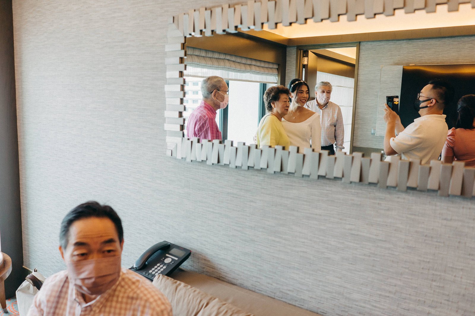 A man in a patterned shirt and mask looks forward in the foreground, while a wedding party is reflected in a mirror, including a bride in a white dress and family members, possibly at a Keyaki Garden Pavilion wedding in Singapore.