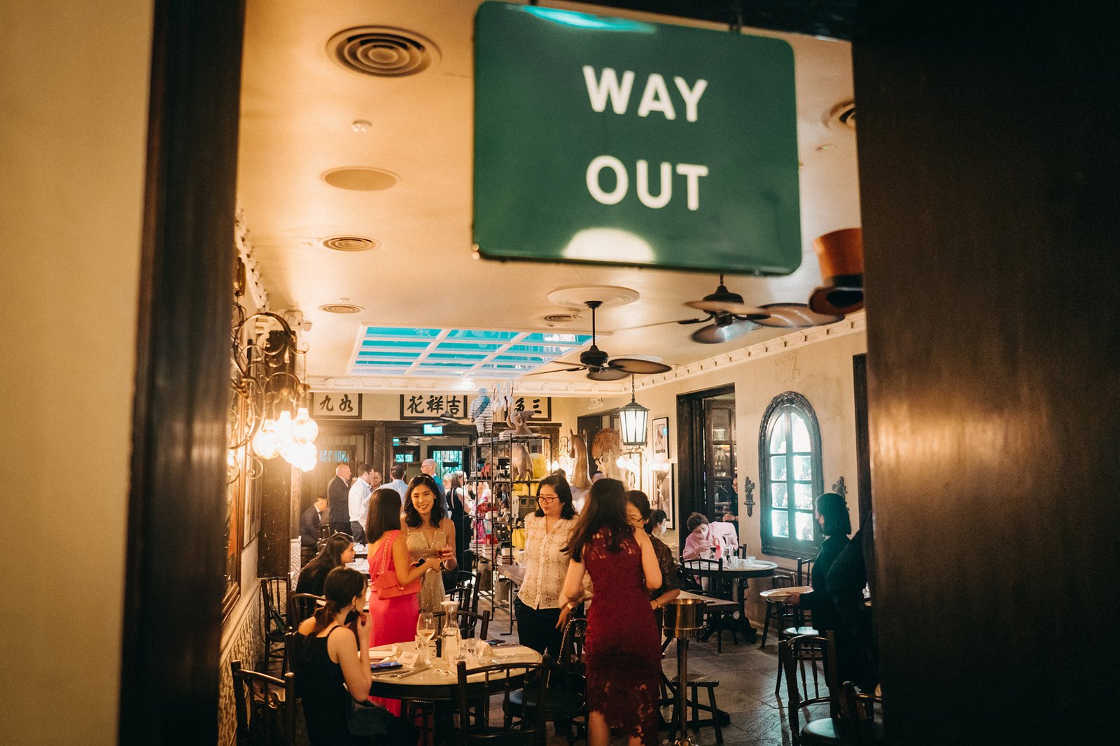 A lively gathering at The English House Singapore, with guests mingling under a "WAY OUT" sign. Warm lighting illuminates the elegant interior, hinting at a wedding celebration.