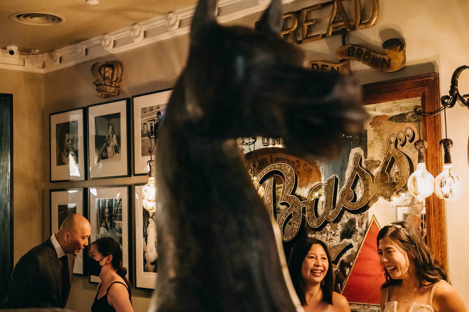 A blurred horse head statue in the foreground at The English House Wedding Singapore, with guests smiling and talking in the background near a vintage mirror with "Bass" lettering.